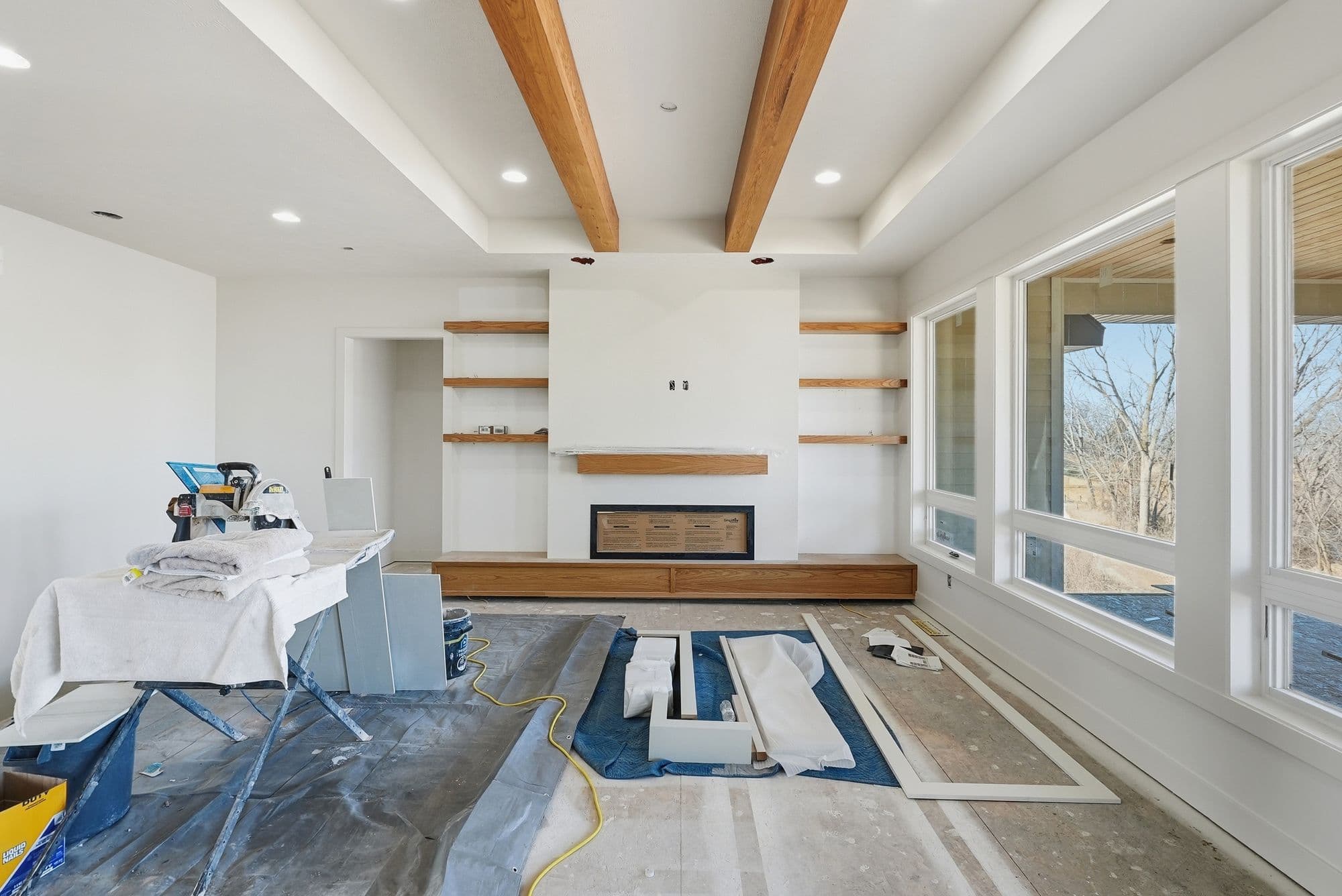 Living room under construction features white walls, hardwood floors covered with protective materials, and wooden ceiling beams. A modern fireplace with built-in shelving sits against the wall, and large windows provide natural light.