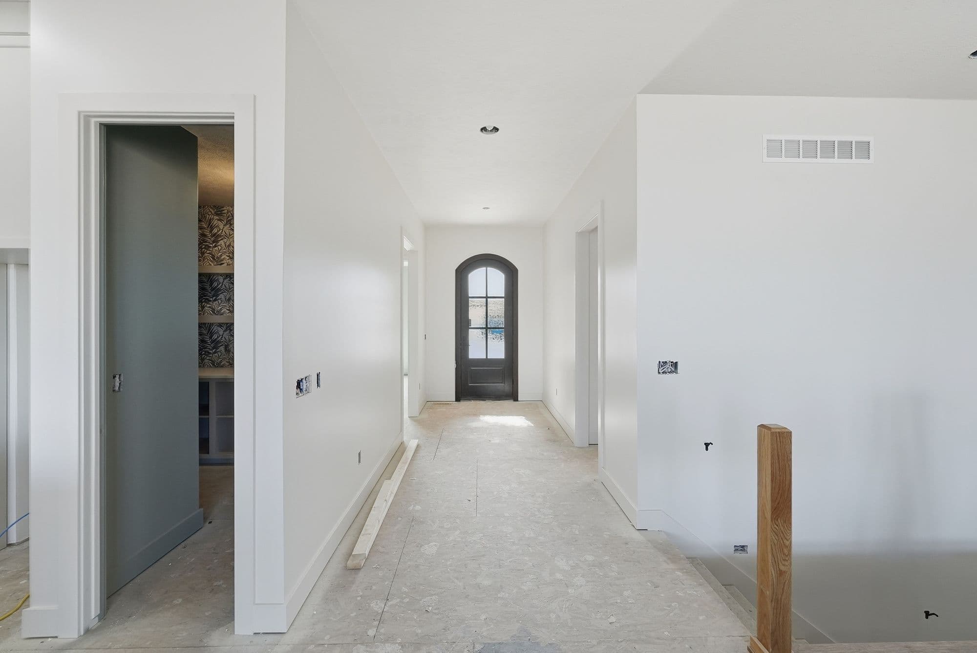 Bright hallway with white walls and trim leads to an arched doorway, with a partially open doorway revealing a room with wallpaper. The unfinished floor is light and the space is being renovated.