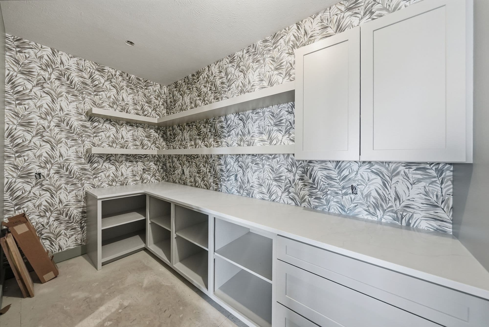 Pantry with gray leaf wallpaper, white cabinets, and white countertops. The pantry also features open shelving and drawers, with a concrete floor.