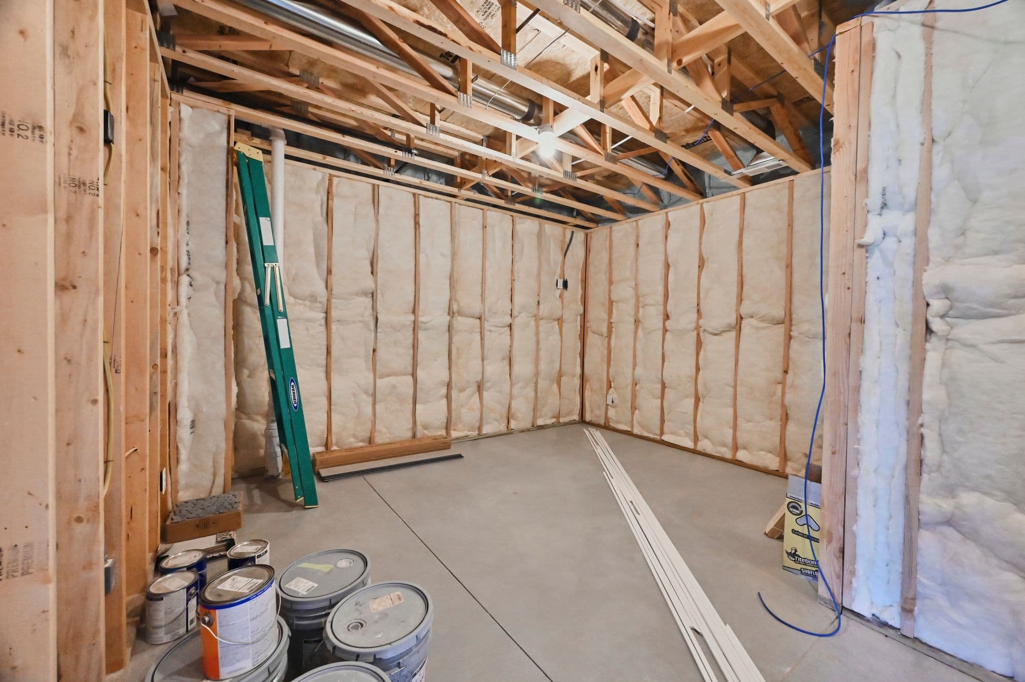 Unfinished basement room with exposed framing and insulation. The room contains construction materials, paint buckets, and a ladder, indicating ongoing work.