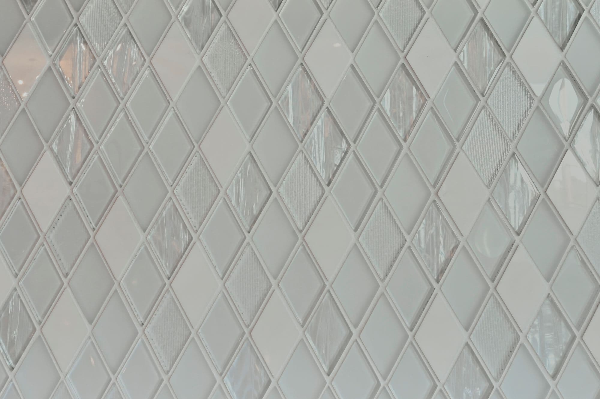 Close up view of diamond-patterned backsplash featuring white and glass tiles with thin grout lines. The tiles have varying textures and finishes, creating a visually interesting surface.