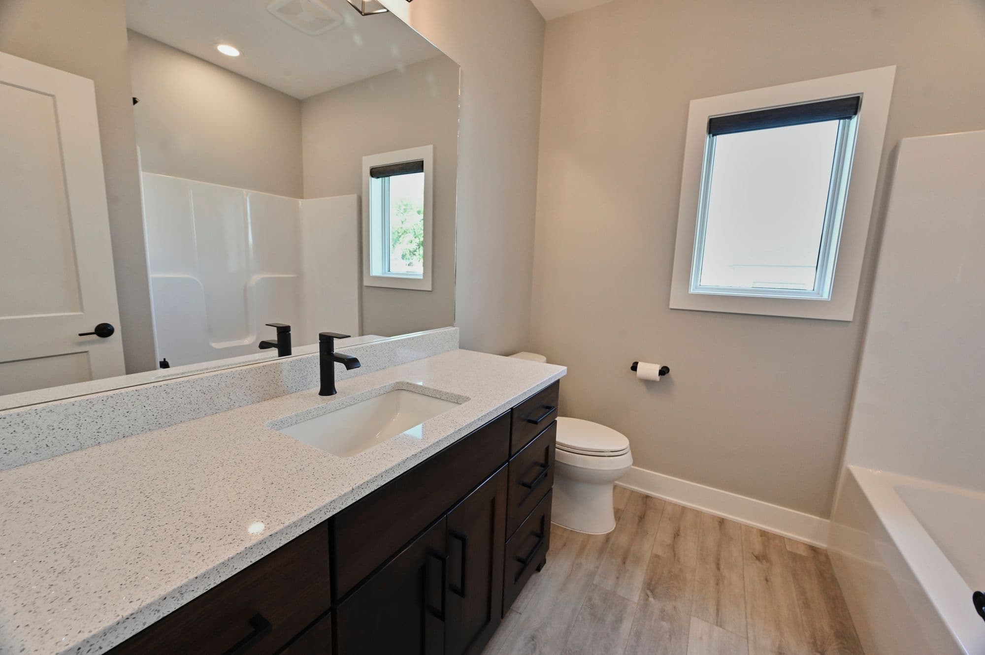 Bathroom with white quartz countertops, dark wood vanity, and light wood flooring. The room features a window with dark shade, white toilet, and a shower/tub.