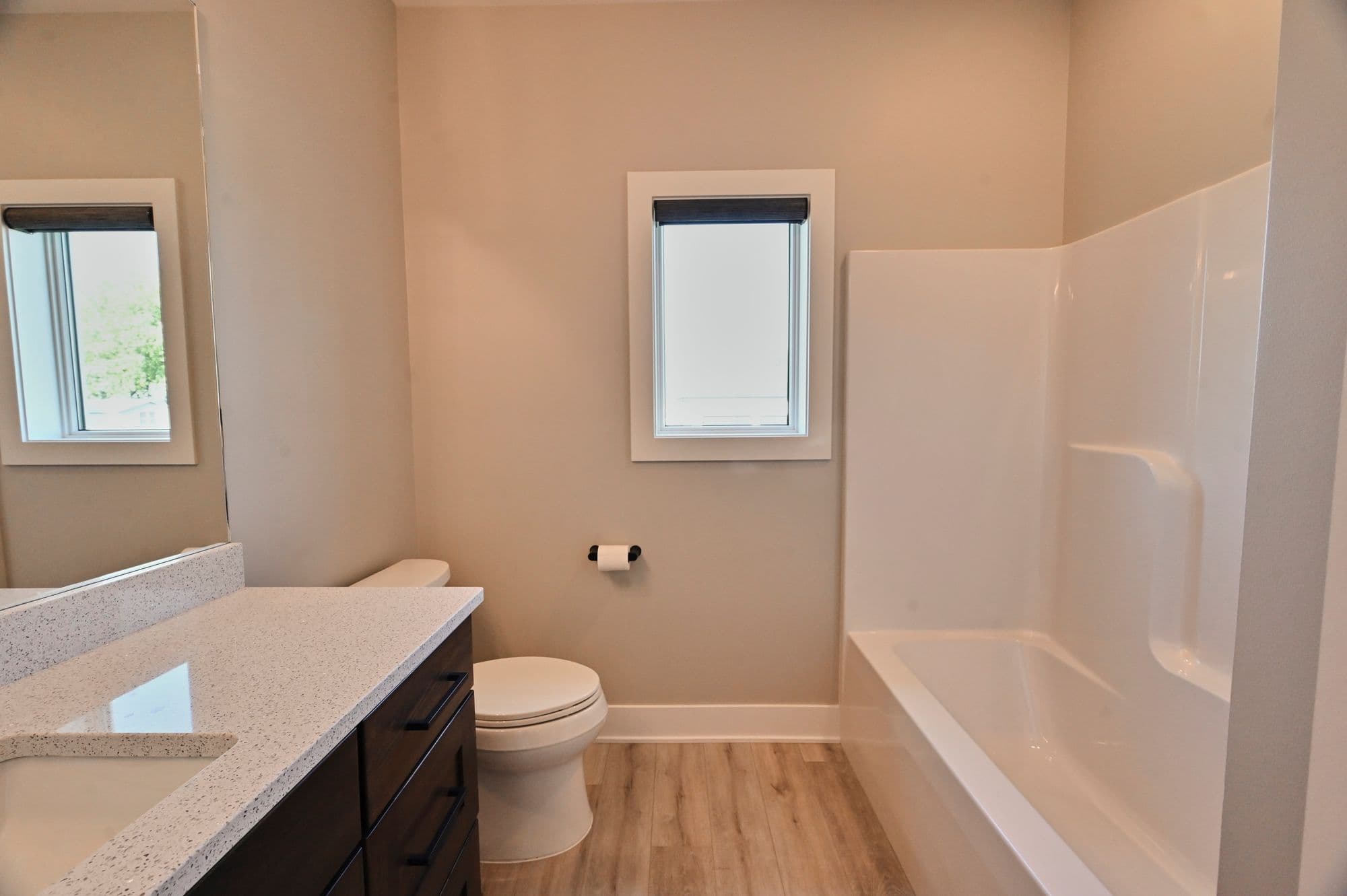 Bright bathroom featuring a dark vanity with a white countertop, a toilet, a white window with a dark shade, and a white bathtub with shower surround. The walls are light beige and the flooring is a light wood-look vinyl.