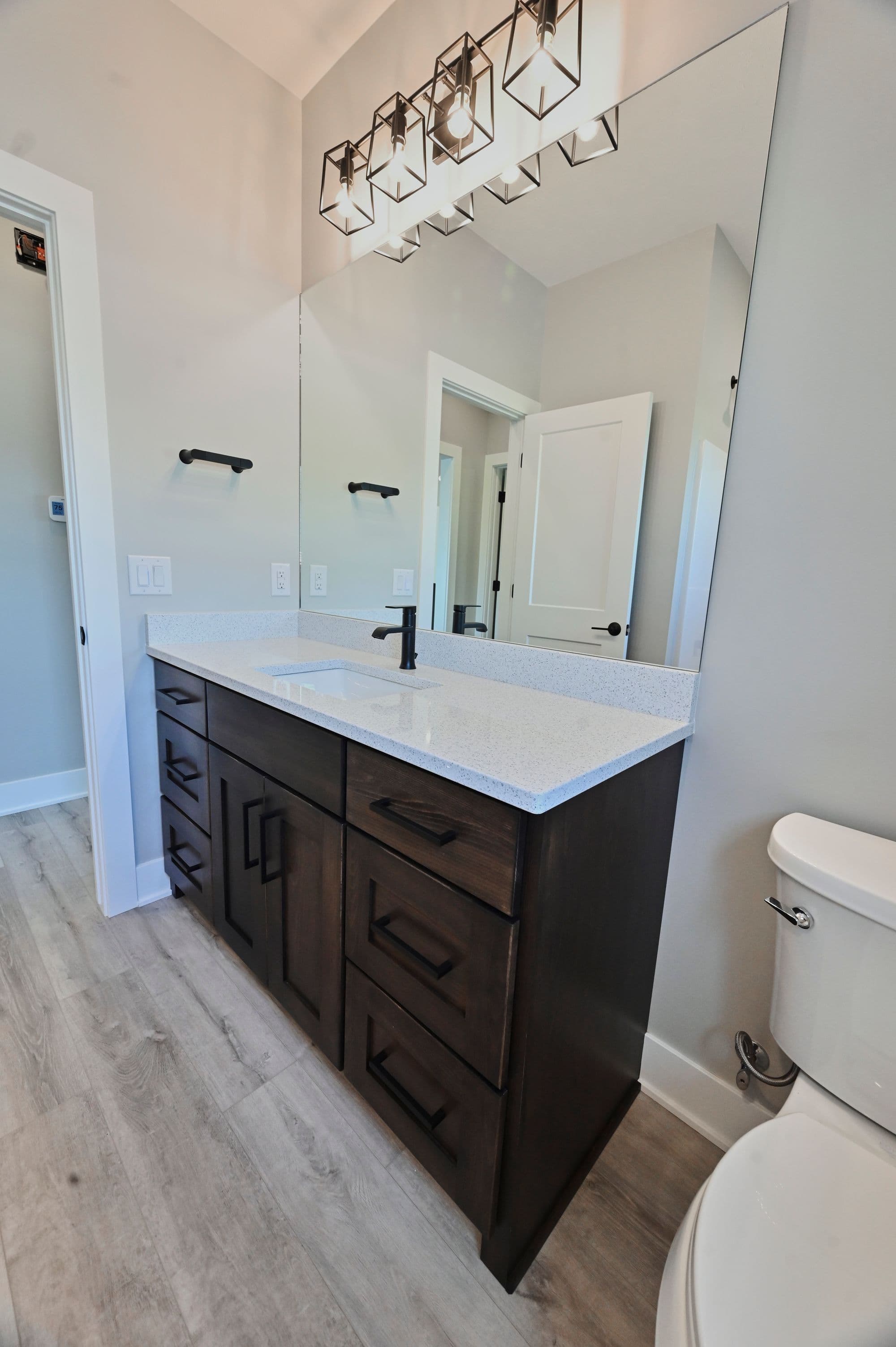 Bathroom features a dark wood vanity with a white countertop and black fixtures, along with a large mirror and modern lighting. The flooring is light gray wood-look vinyl.