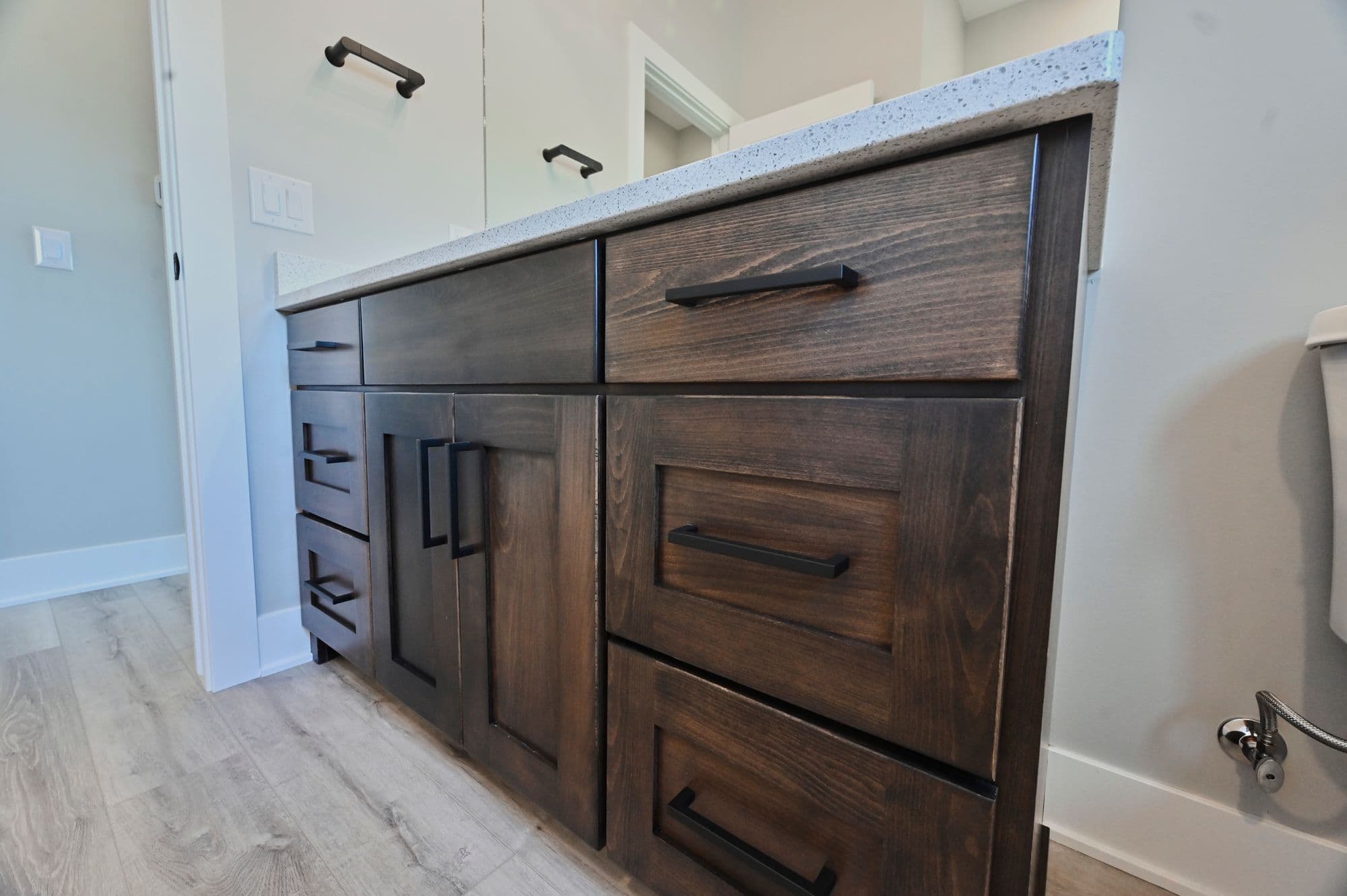This bathroom features a custom wood vanity with dark brown stain, complemented by a white countertop with subtle speckles. Matte black hardware on the cabinets and drawers adds a modern touch.