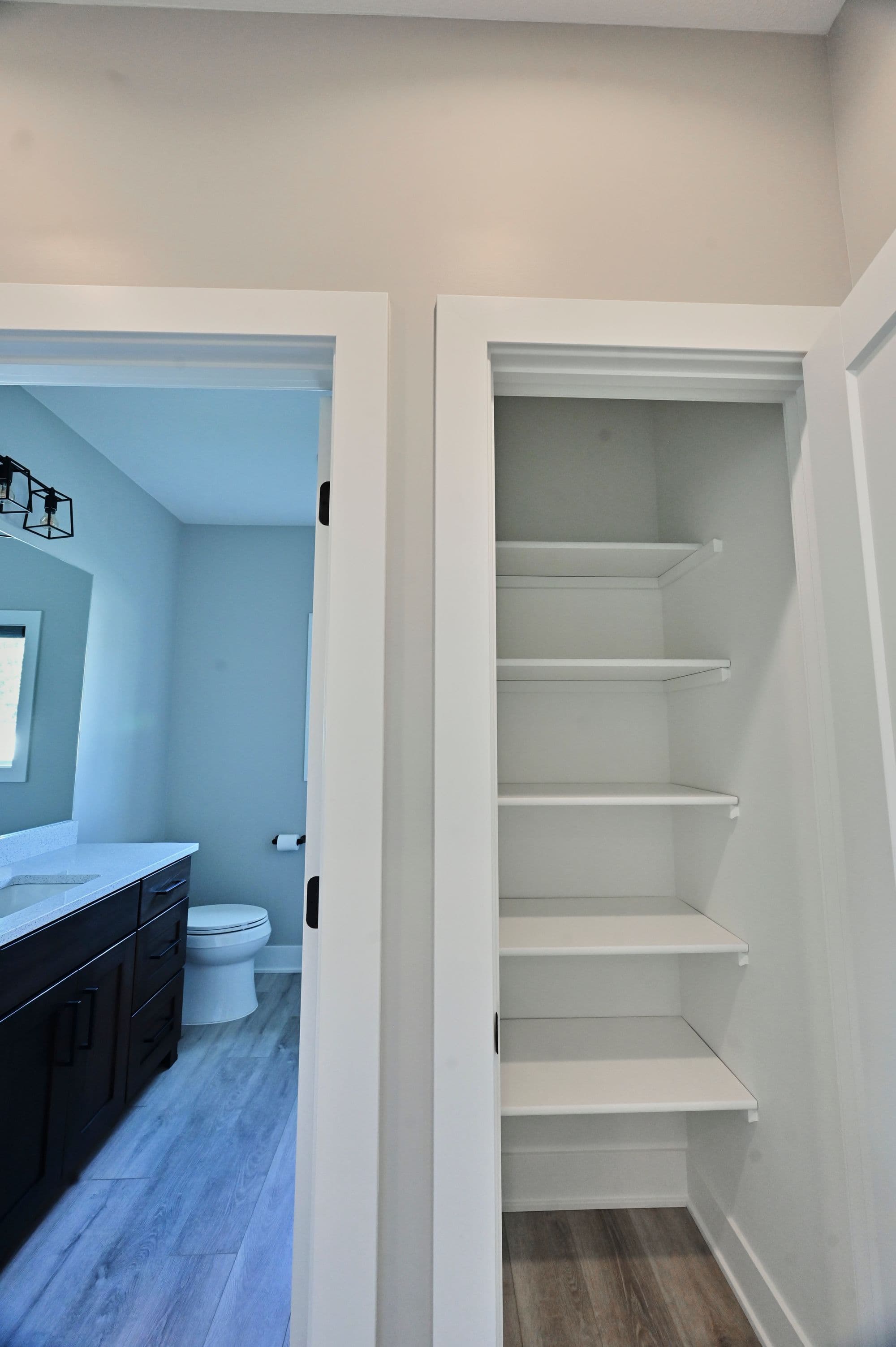 Bright bathroom and adjacent linen closet with white shelving in a modern home. The bathroom features a dark wood vanity, white countertops, toilet and gray plank flooring.