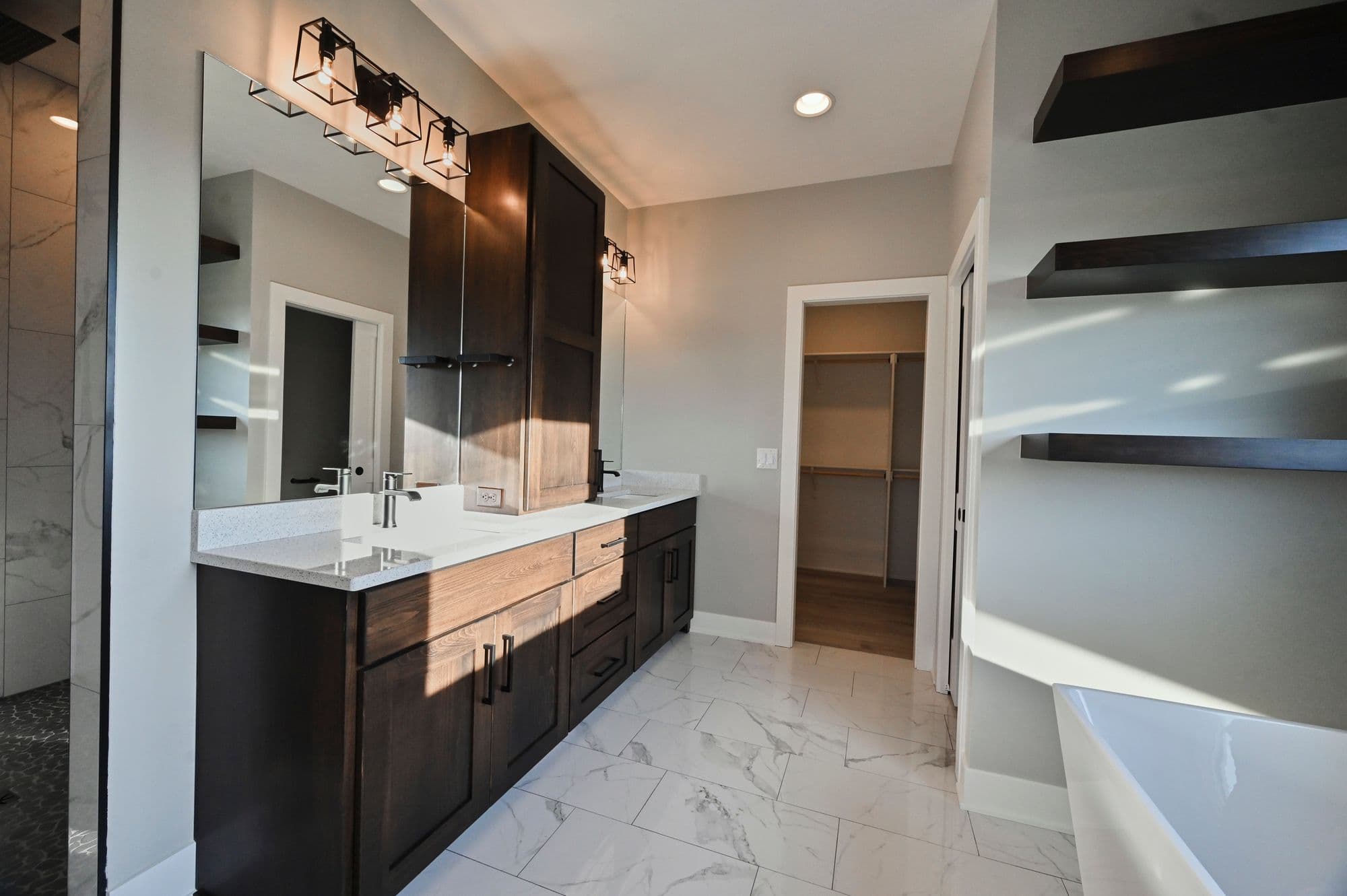 Modern bathroom featuring a double vanity with dark wood cabinets and white countertops under caged sconces, a large mirror, and a walk-in closet. The floor is tiled with marble-patterned tiles and the walls are painted a soft gray.