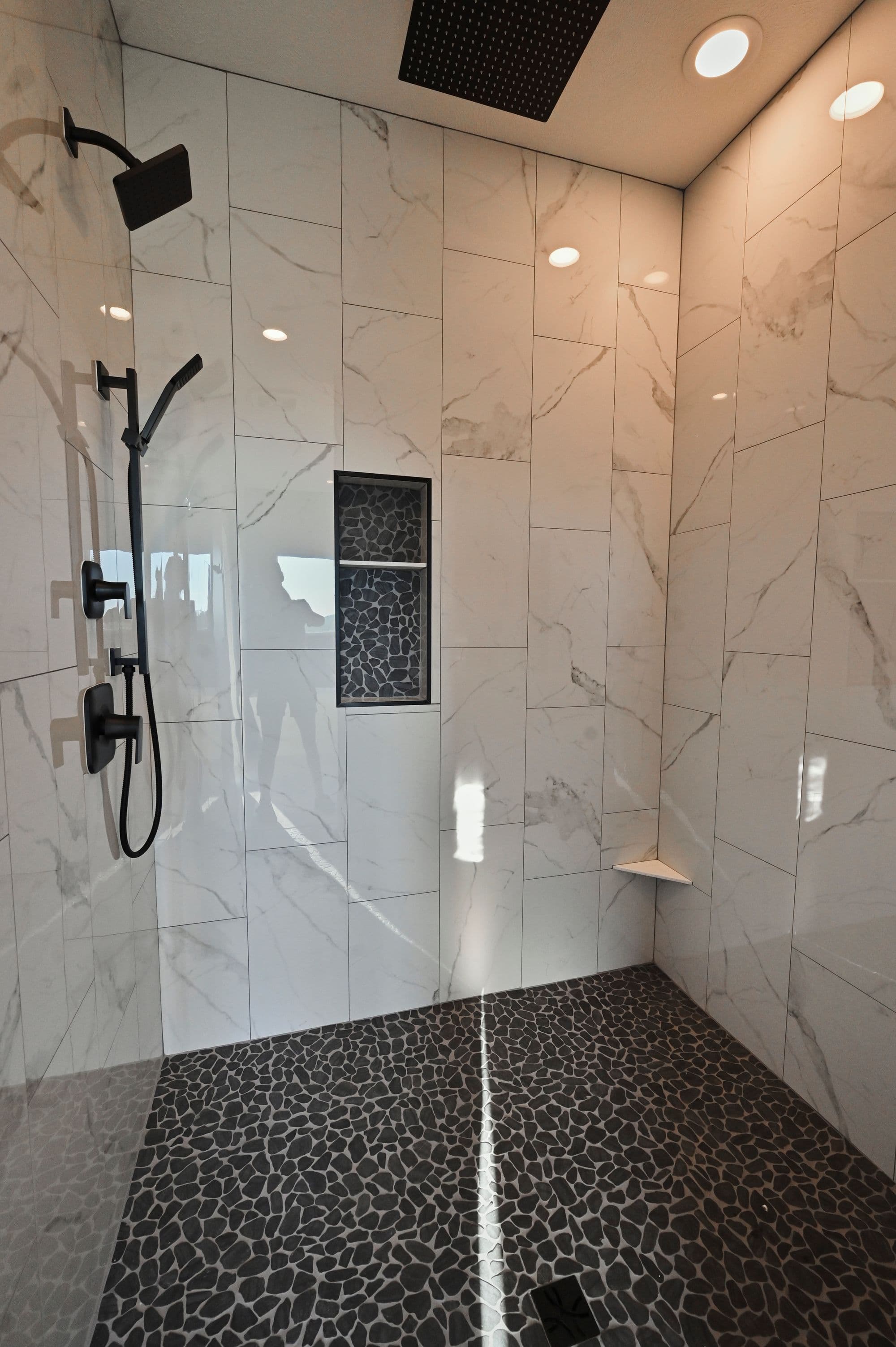Modern bathroom featuring large-format white marble tiles, a black pebble stone floor, and contemporary black fixtures including a rainfall shower head and hand shower. A built-in niche with matching black pebble stones adds a design accent.