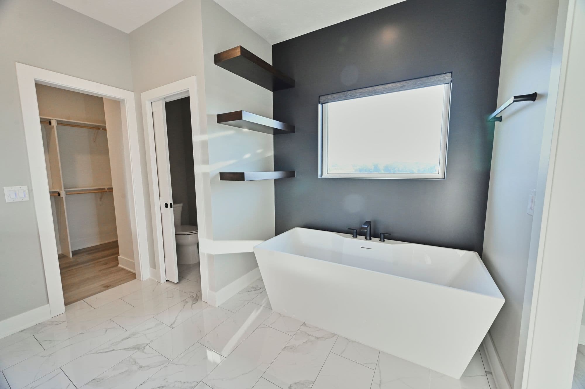 Modern bathroom features a freestanding white tub under a window, contrasting against a dark gray accent wall. Three floating wooden shelves are mounted on the adjacent light gray wall.