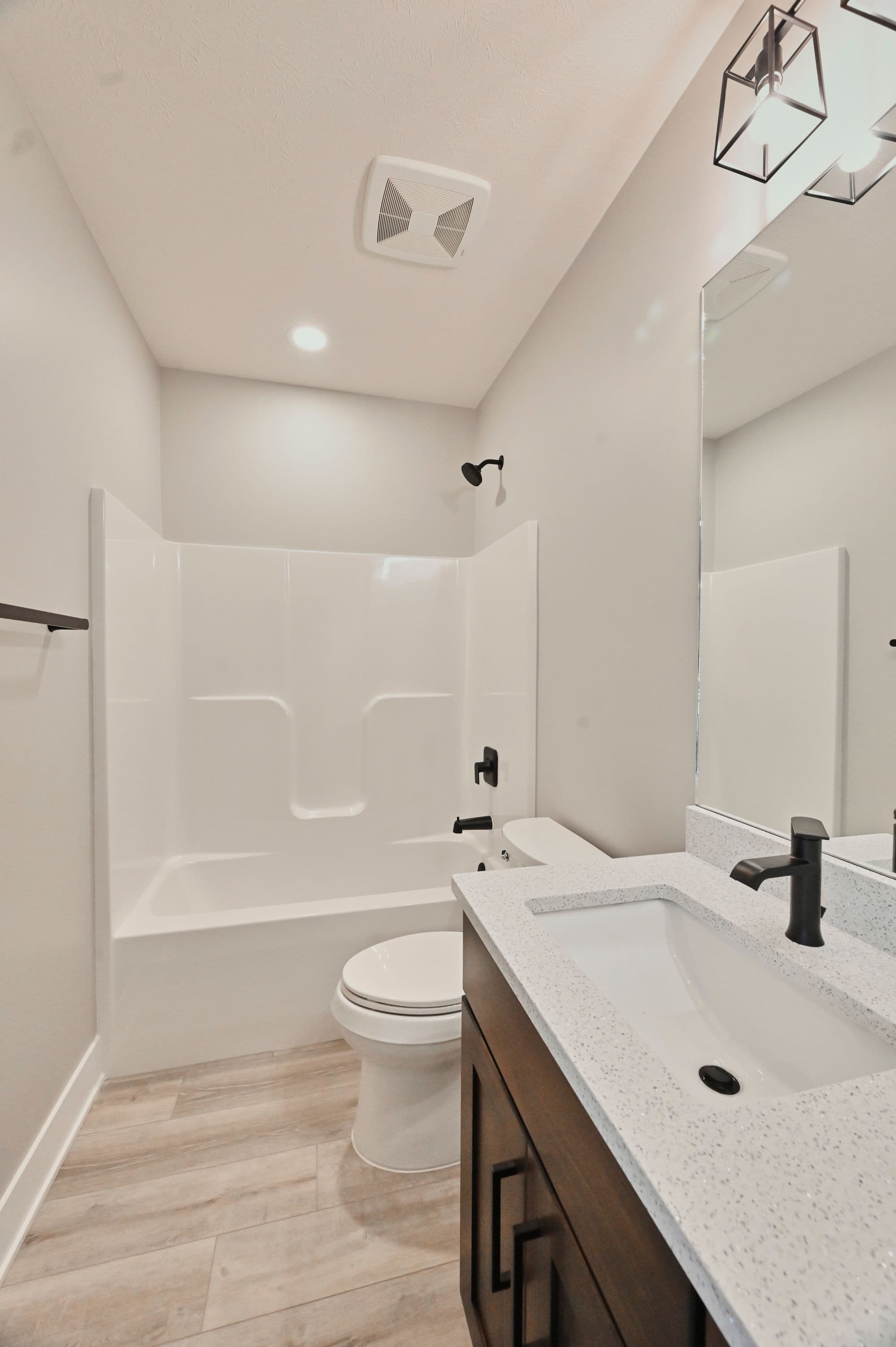 Bathroom with light wood flooring, white shower and tub, and dark brown vanity cabinet topped with white speckled countertop. The room also includes a white toilet and a framed vanity mirror.