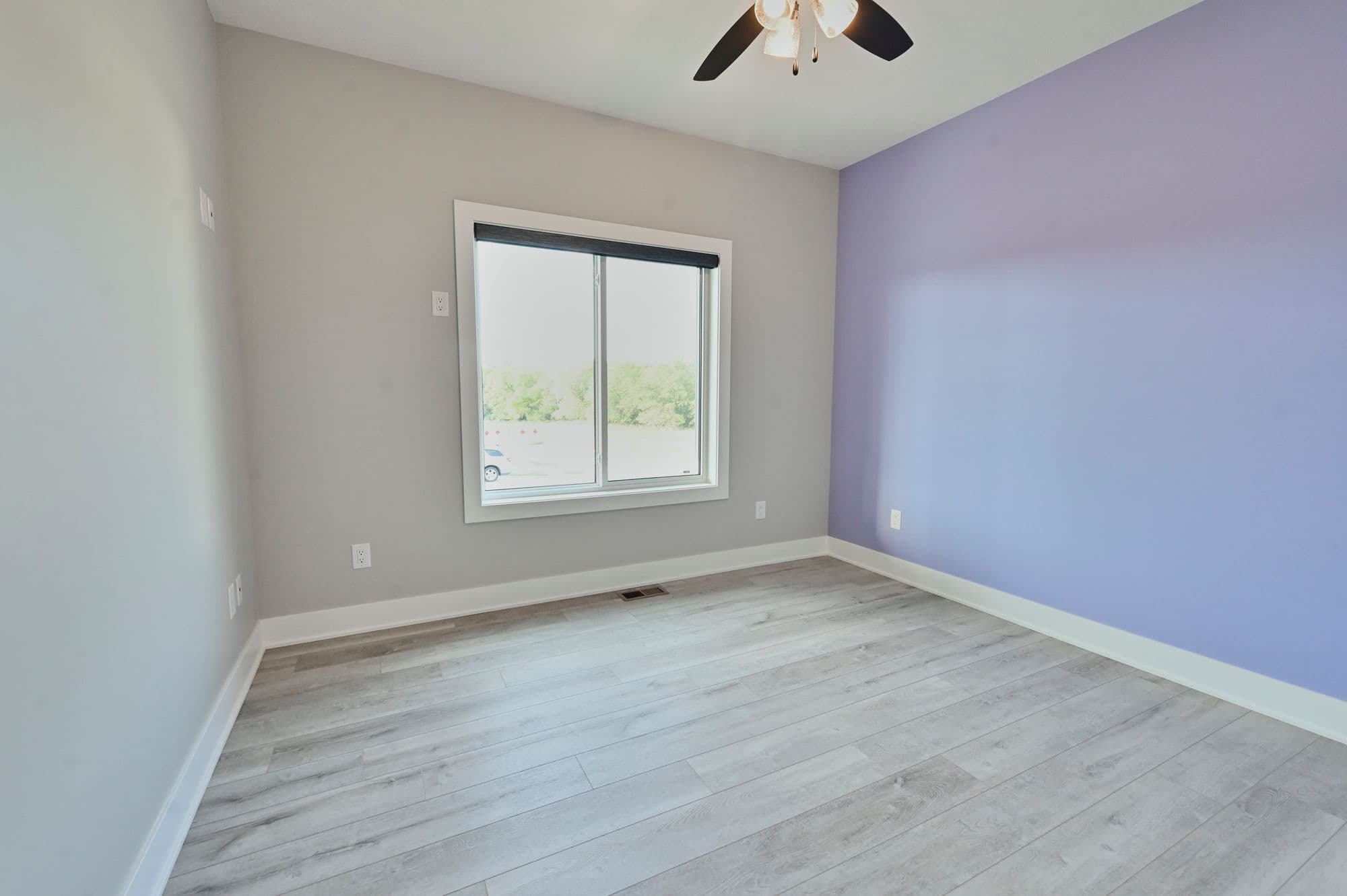 An interior bedroom features light gray walls and a single window, contrasting with a lilac-painted accent wall. The floor is light gray wood-look planking, and a ceiling fan is visible.