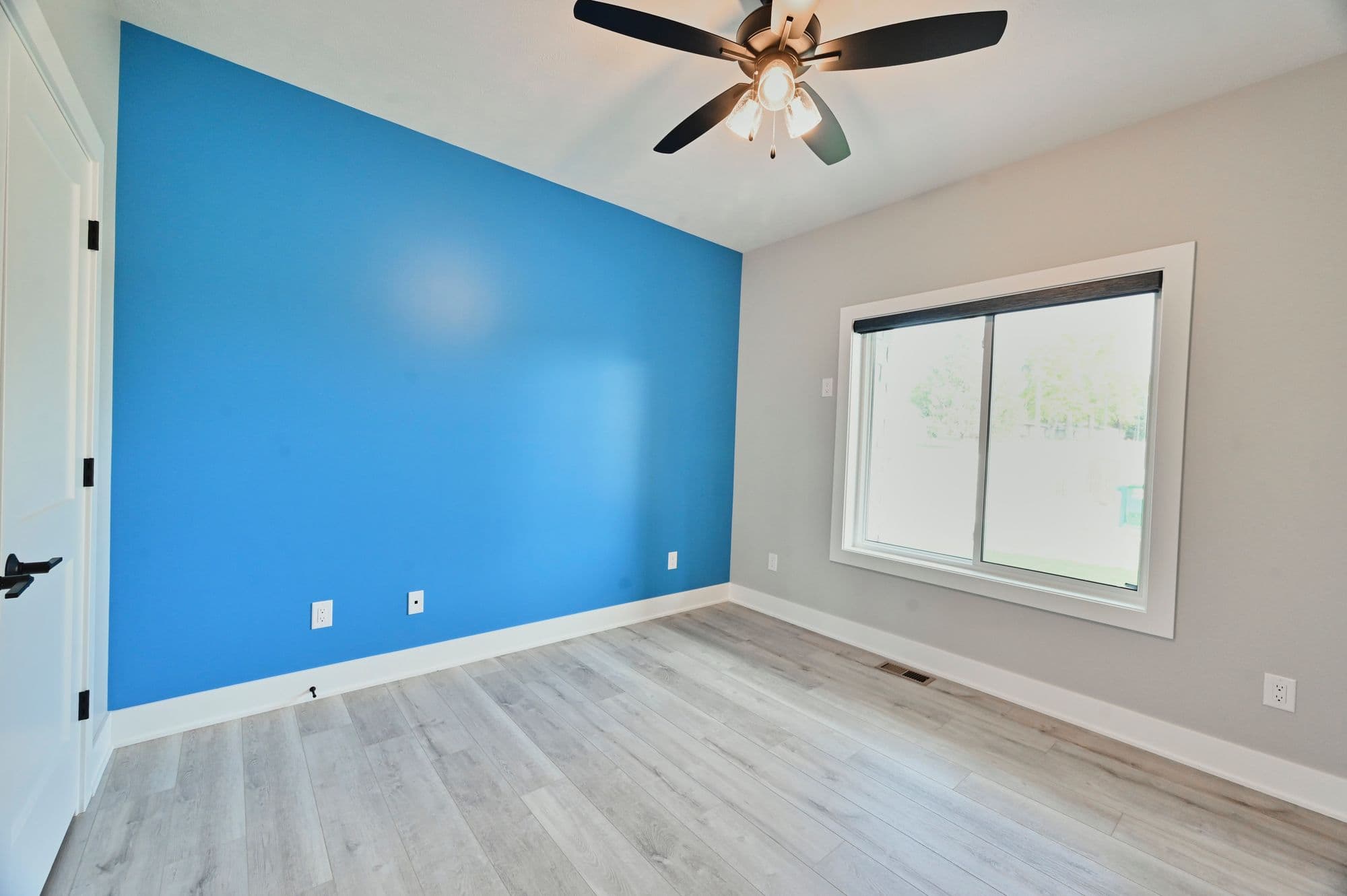 A bedroom with a blue accent wall, light gray walls and laminate floors. A window lets natural light into the room, which is also illuminated by a ceiling fan.