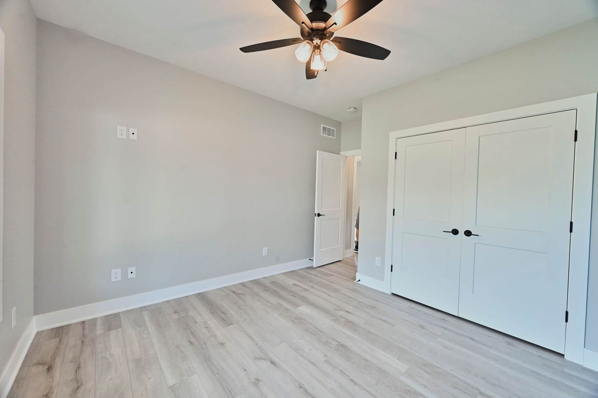 Bright bedroom with light gray walls and light wood-look flooring, featuring a ceiling fan and a closet with white paneled doors and black hardware. An open doorway leads to another room.