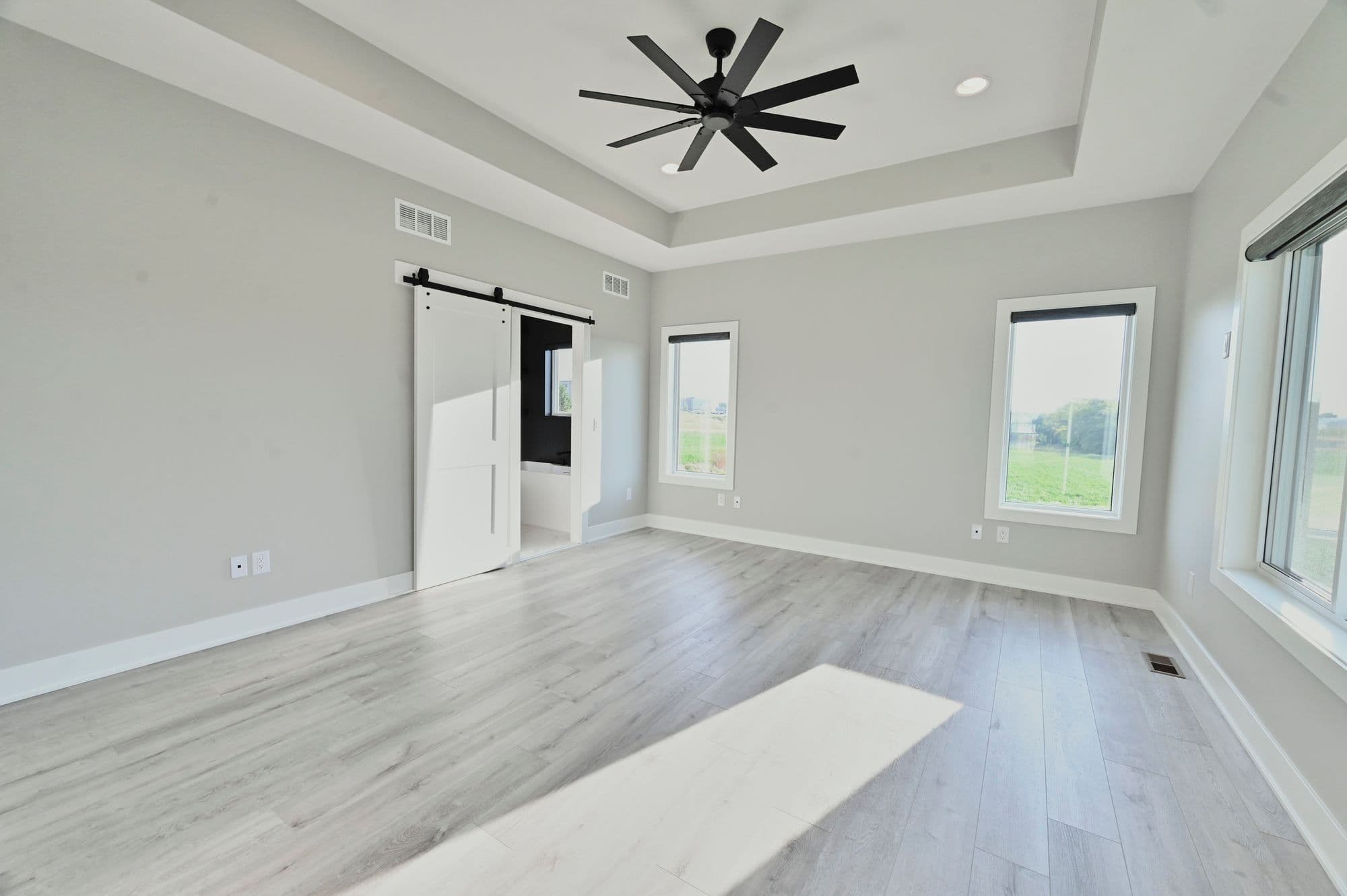 Bright primary bedroom features gray walls, light wood flooring, a black ceiling fan, and two windows with exterior views. A white barn door provides access to the ensuite bathroom.