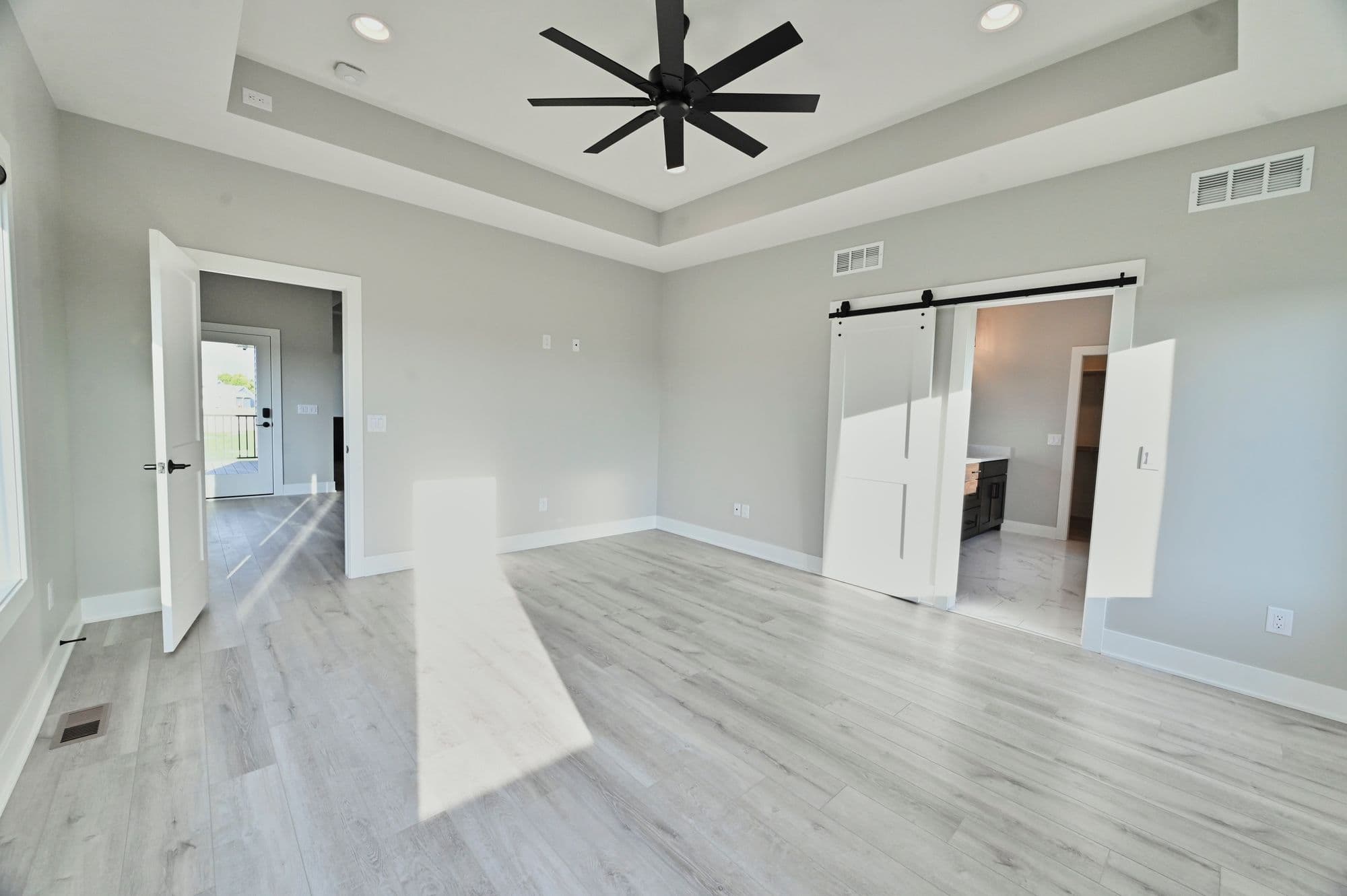 Bright and airy bedroom with light gray walls and light wood-look floors. A modern ceiling fan is centered, and open doorways and sliding barn doors lead to other rooms.