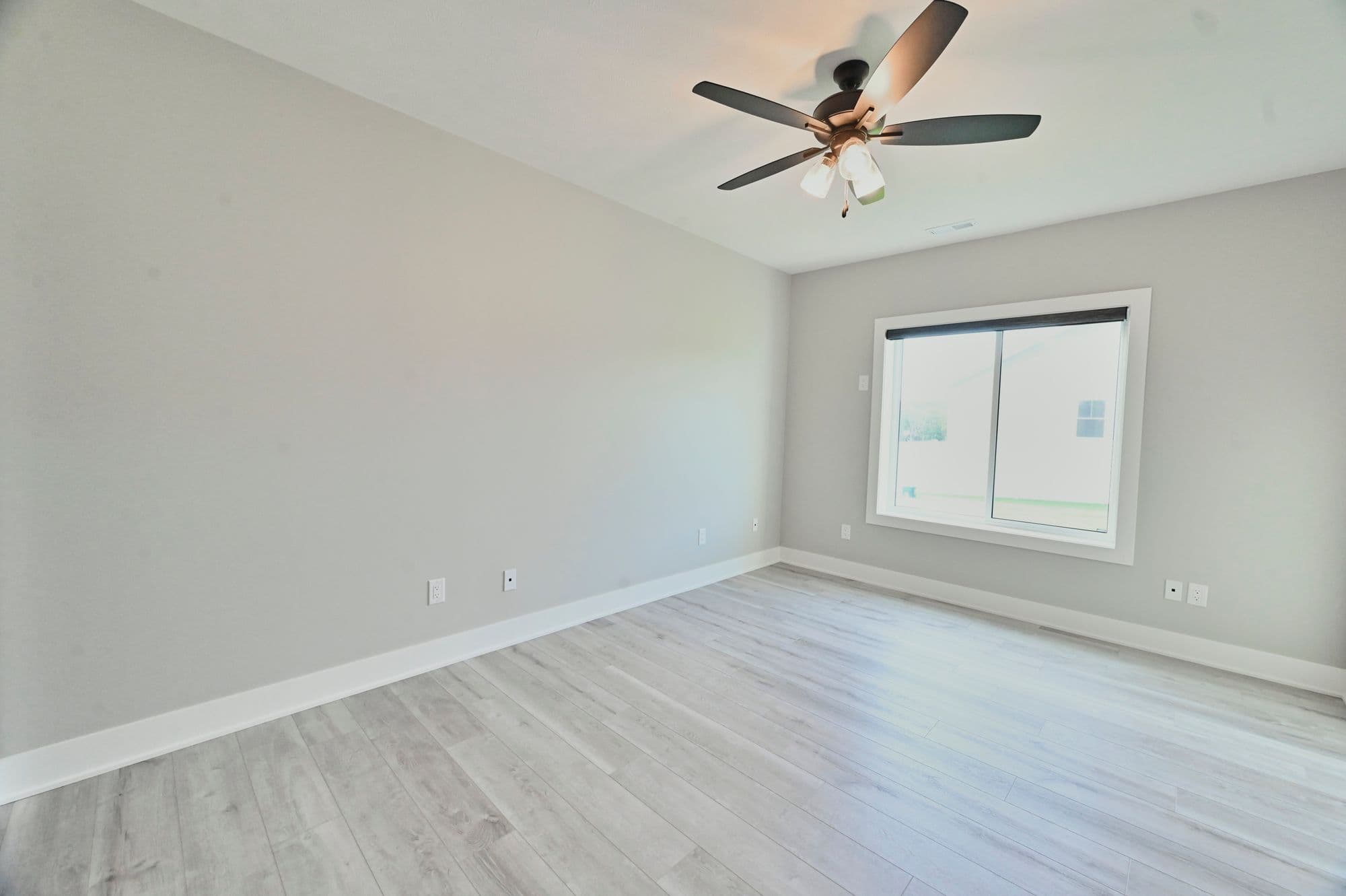 This is a bright empty bedroom featuring light gray walls and wood floors, with a modern ceiling fan and a window that lets in natural light. The room is simple and clean with white trim.