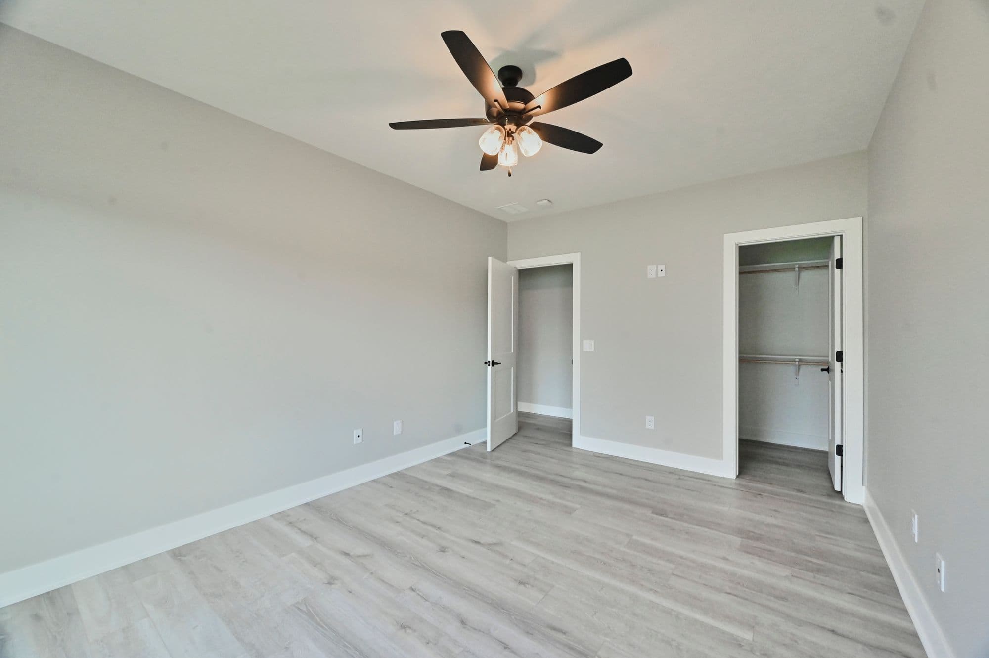 Bedroom with light gray walls and light wood-look flooring features a dark ceiling fan with light fixtures. Two doorways lead to other rooms or closets.
