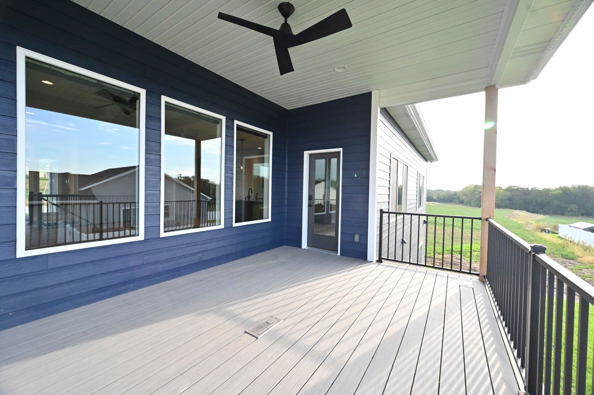 Covered deck features blue siding, a light gray composite deck, and a black ceiling fan. White trim surrounds the windows and door to the interior of the house.
