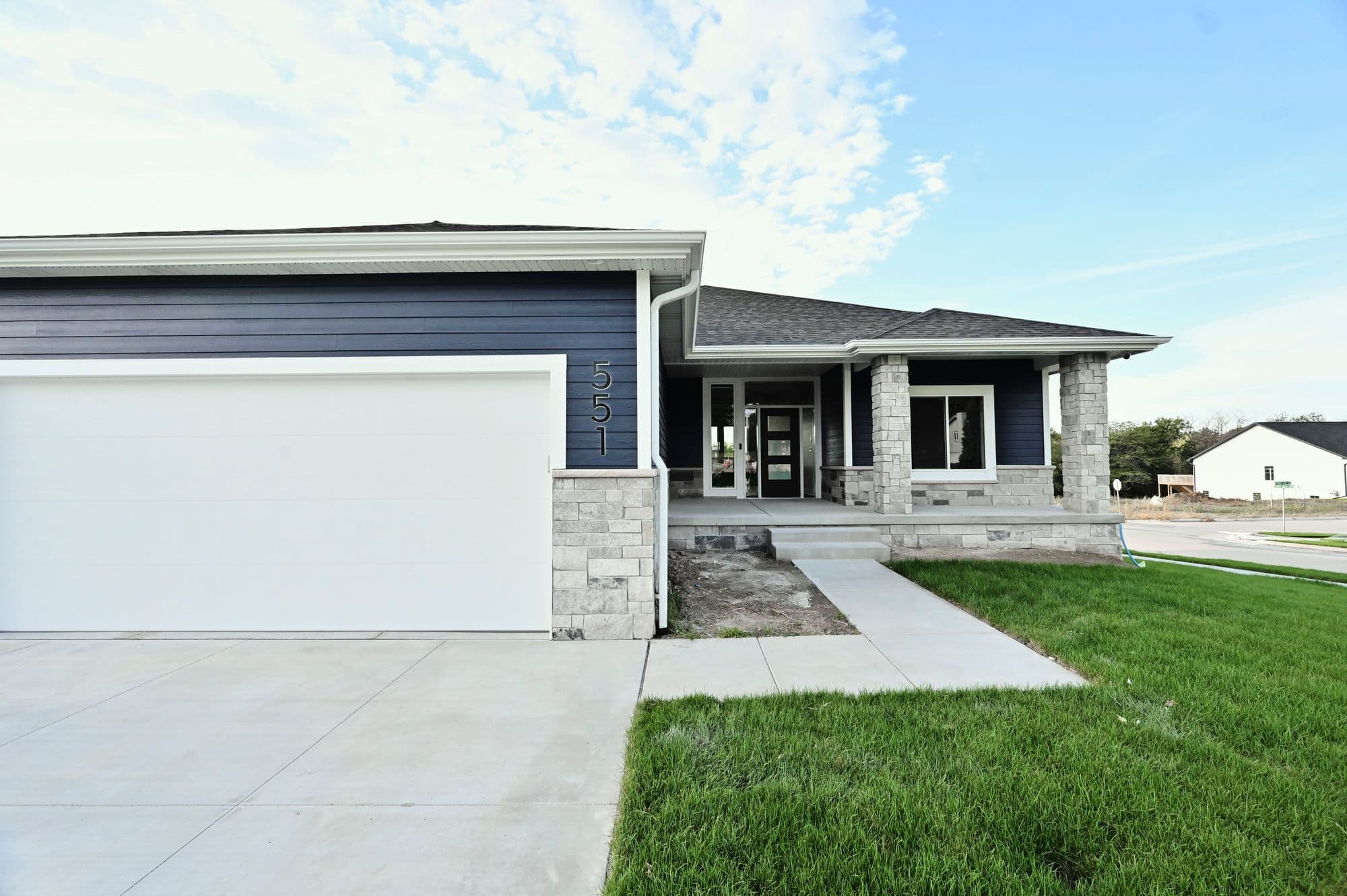 Exterior view of a modern home with a light gray concrete driveway and walkway leading to the entrance. The house features dark blue horizontal siding, stone veneer accents, a white garage door, and the house number '551' affixed to the wall.