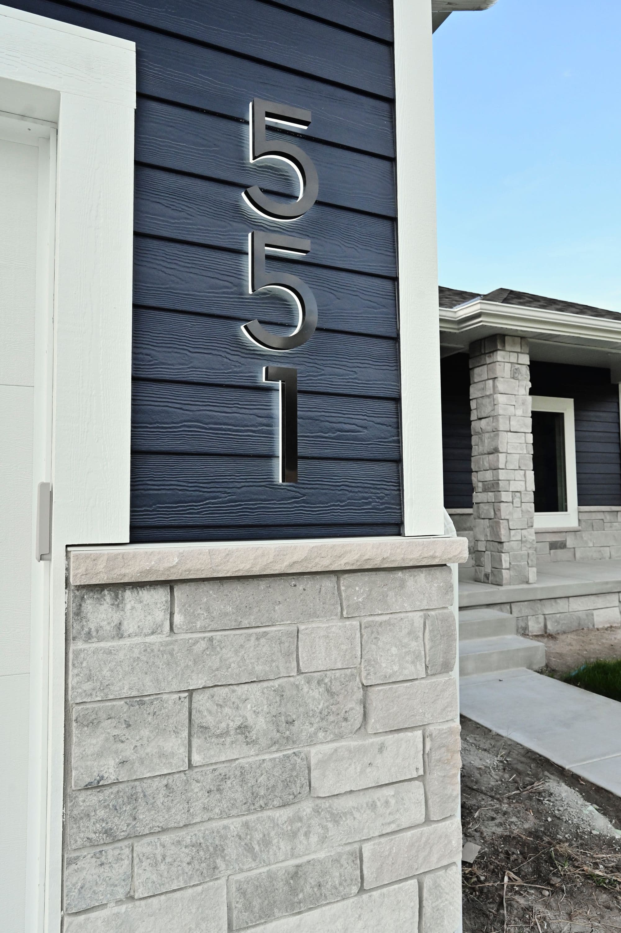 Exterior view of a modern home featuring a blue horizontal siding, stone veneer accents, and a prominent house number display with metallic numerals. The facade includes a white door frame and a section of a stone-clad porch pillar, with a glimpse of the home's entrance in the background.