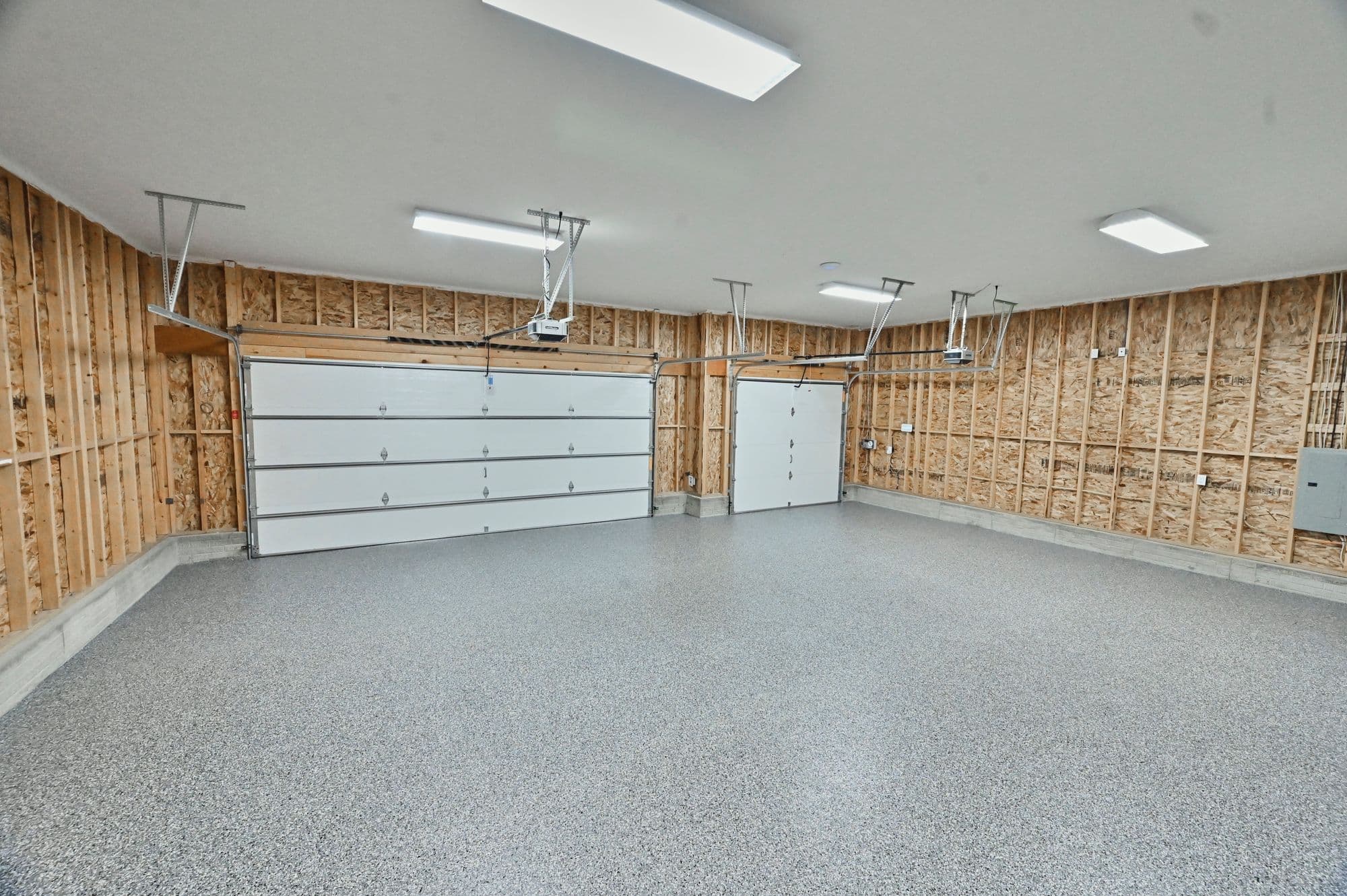 Spacious garage featuring epoxy flooring and unfinished walls with wood framing. Two white garage doors are installed, one for vehicle access and another for pedestrian entry.