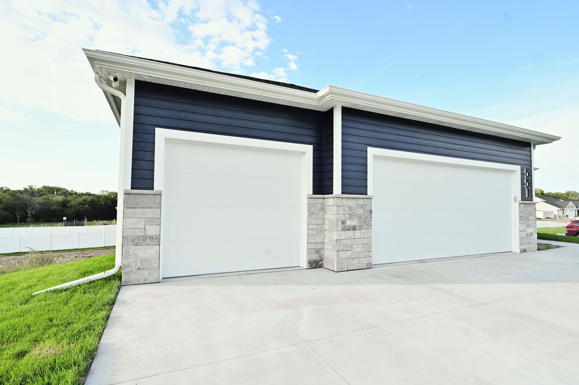 Two-car garage with white doors and stone-veneer pillars. The exterior features navy siding and a white trim, with a concrete driveway.