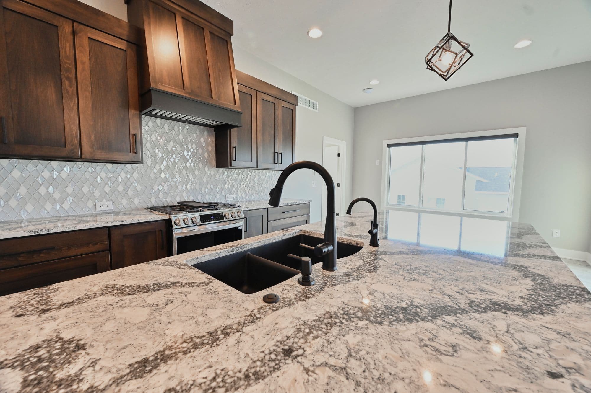 Modern kitchen features dark wood cabinetry, white diamond patterned backsplash and granite countertops. A large island with a double basin sink, black faucet and soap dispenser is centered in the space.