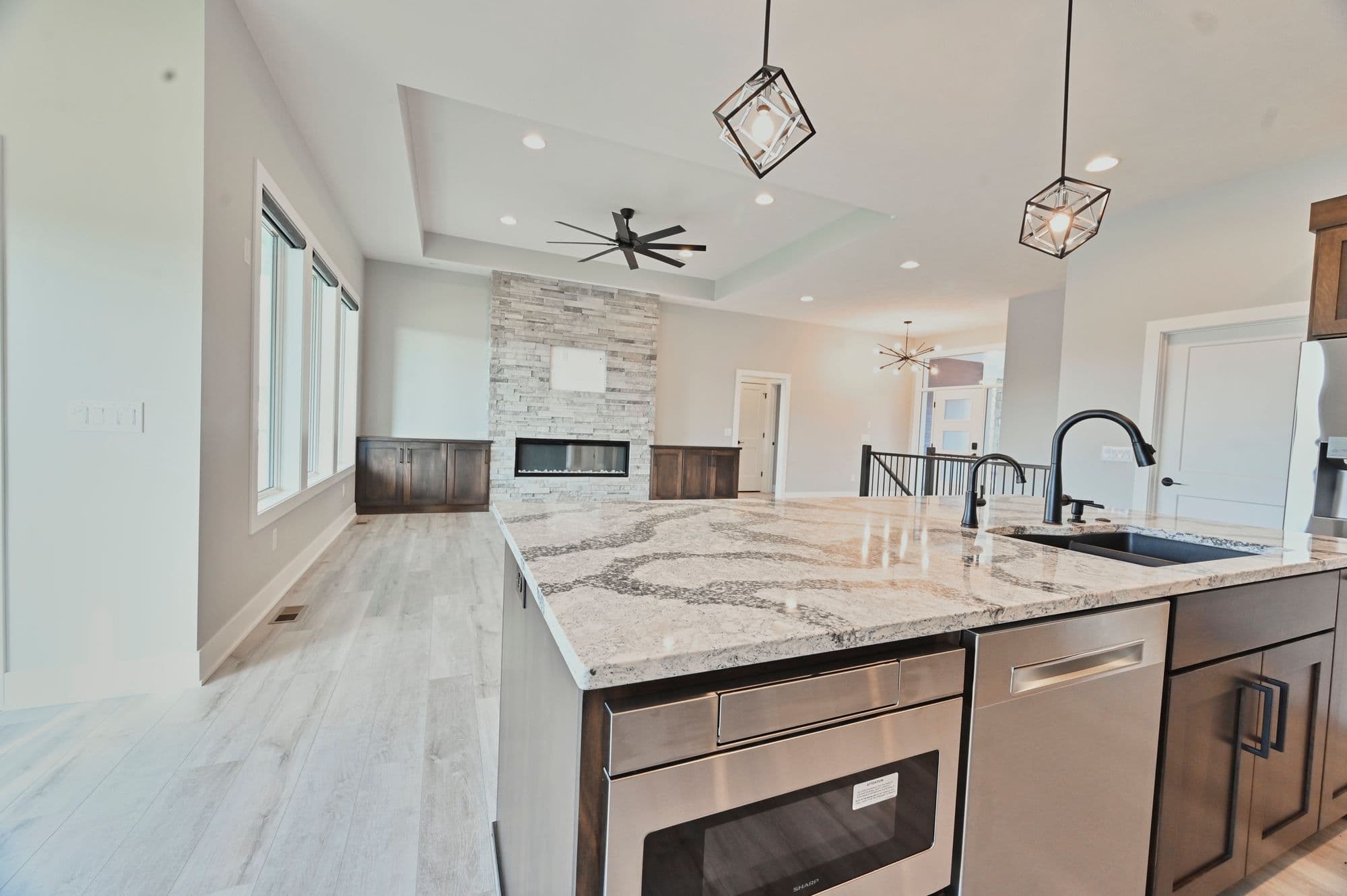 Open-concept living space featuring a modern kitchen island with granite countertops and stainless steel appliances. The living room area has a stone fireplace and neutral-toned decor.