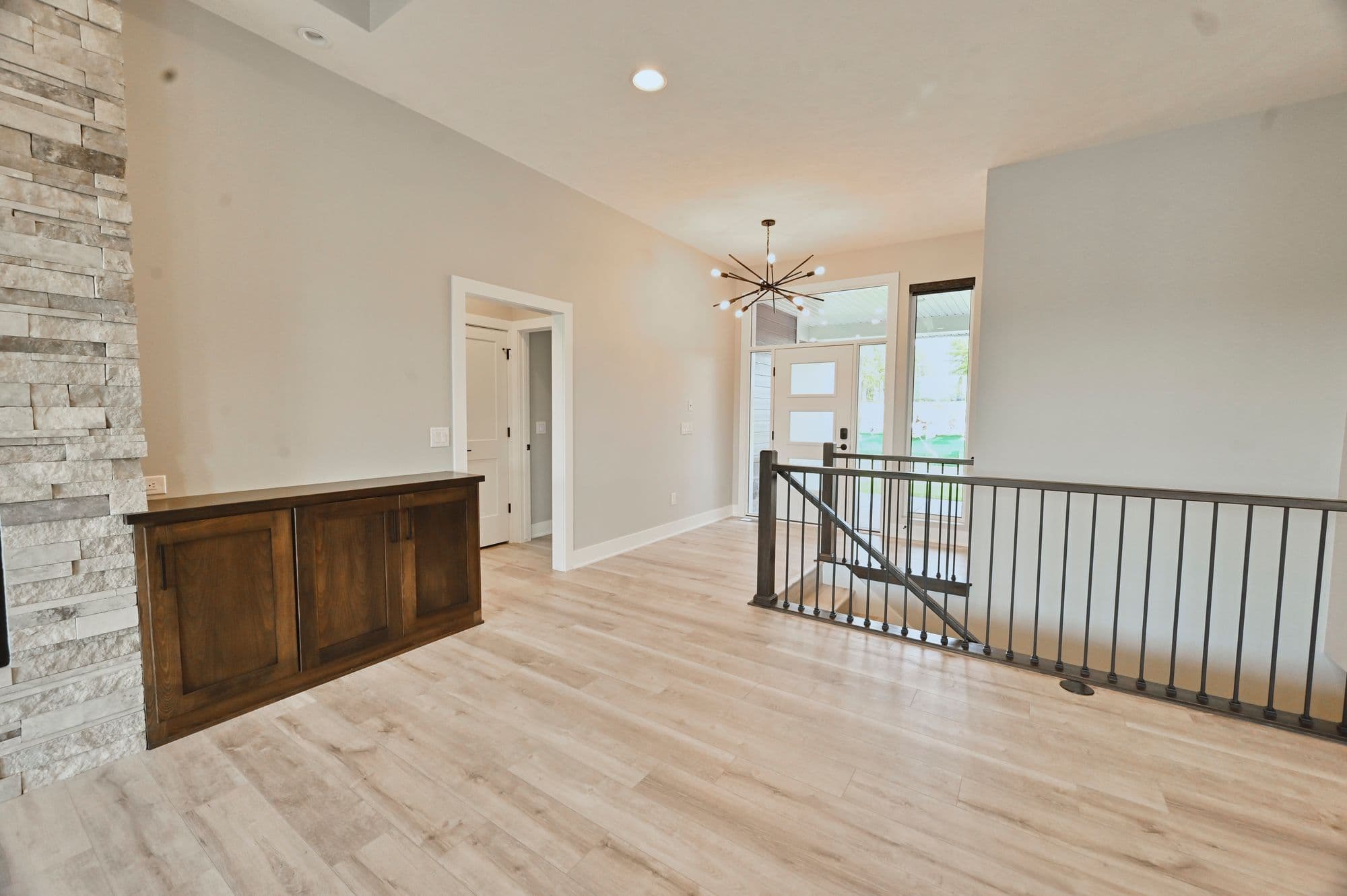 Open-concept living room with light wood floors, stacked stone fireplace, and dark wood built-in cabinets. Black metal railing separates the living room from the entryway, which features a modern chandelier and white front door.