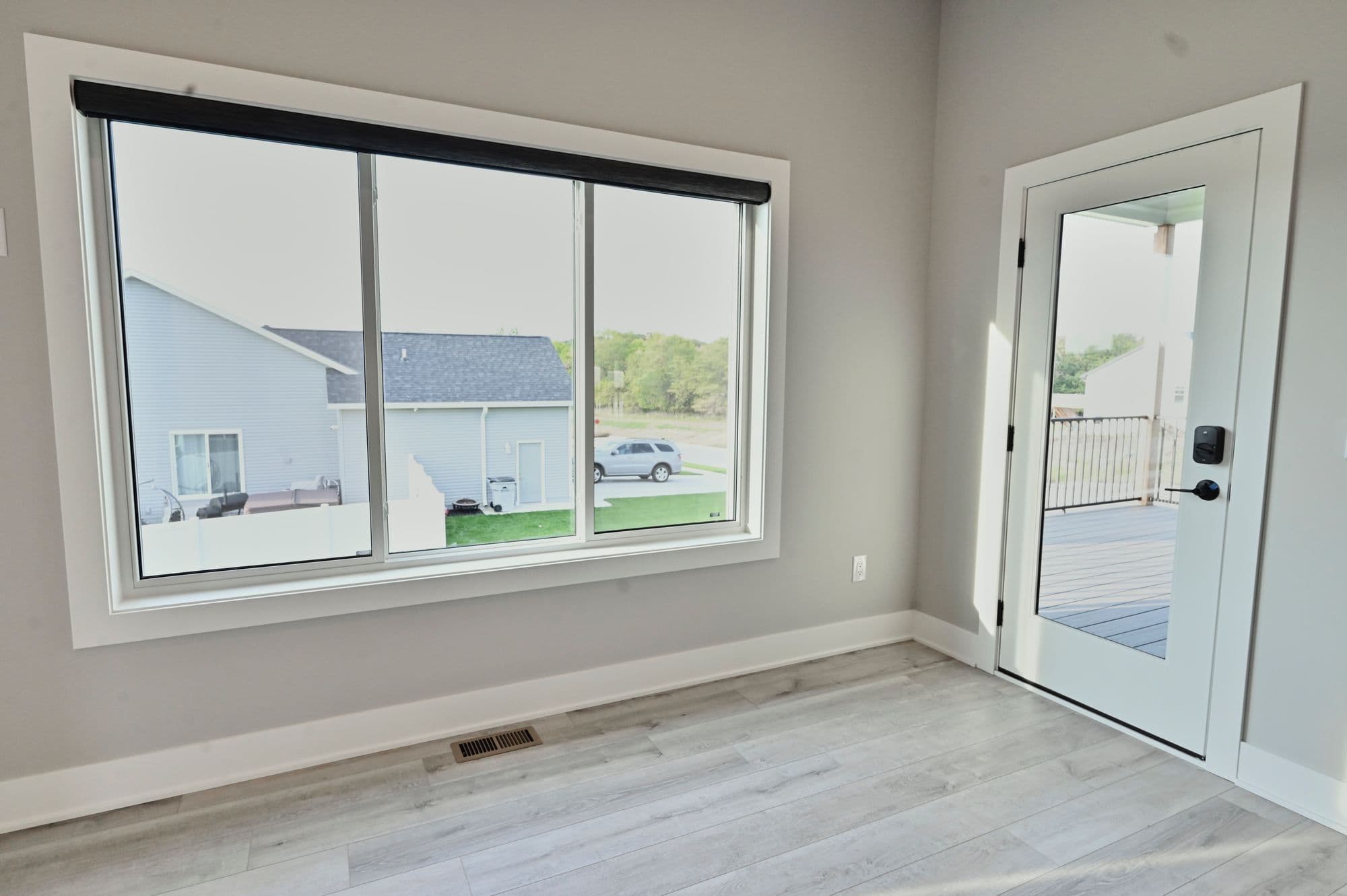 Interior room with light gray walls and light wood-look floors. A large window looks out onto a neighboring house and a door leads to an outdoor space.