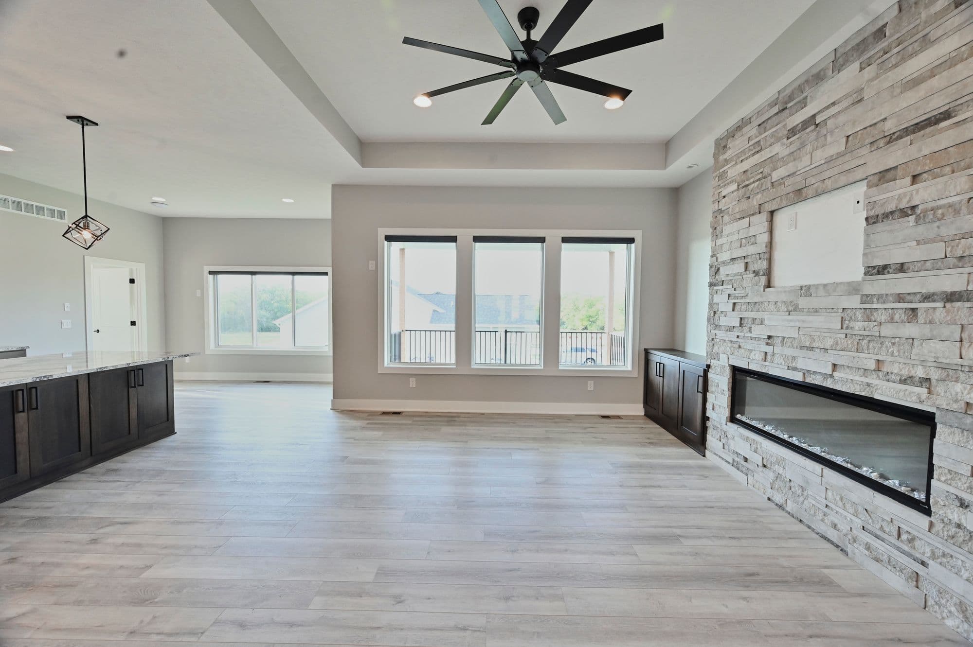 Bright living room features stone accent wall with built-in fireplace and recessed TV space, complemented by modern ceiling fan. Large windows provide ample natural light, and dark wood cabinetry adds contrast to light wood floors.