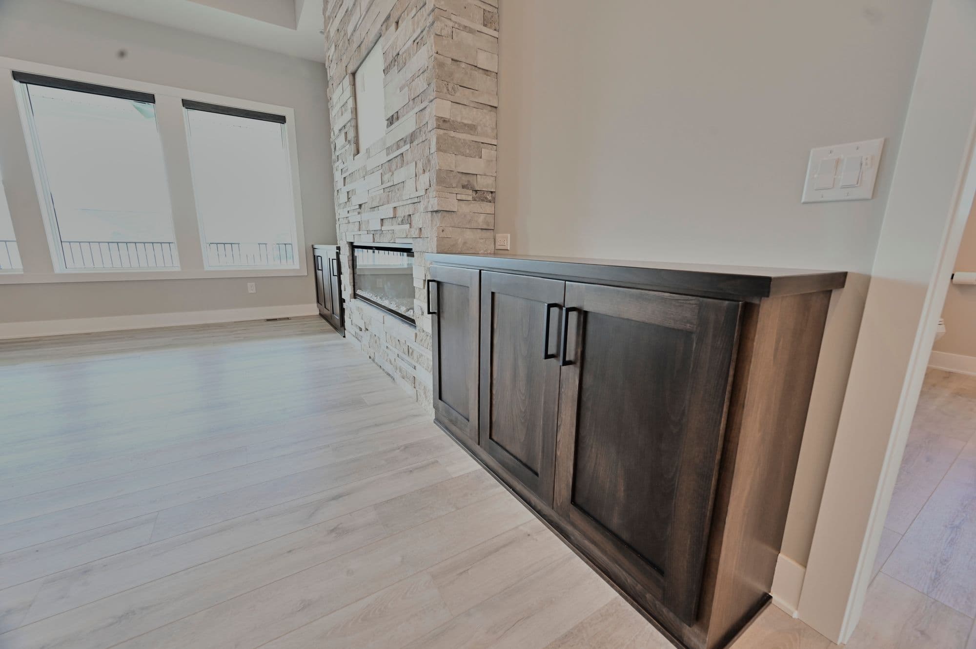 Contemporary living room features a stone fireplace with built-in cabinets, light wood flooring, and large windows. The cabinets are a dark stained wood with black hardware.