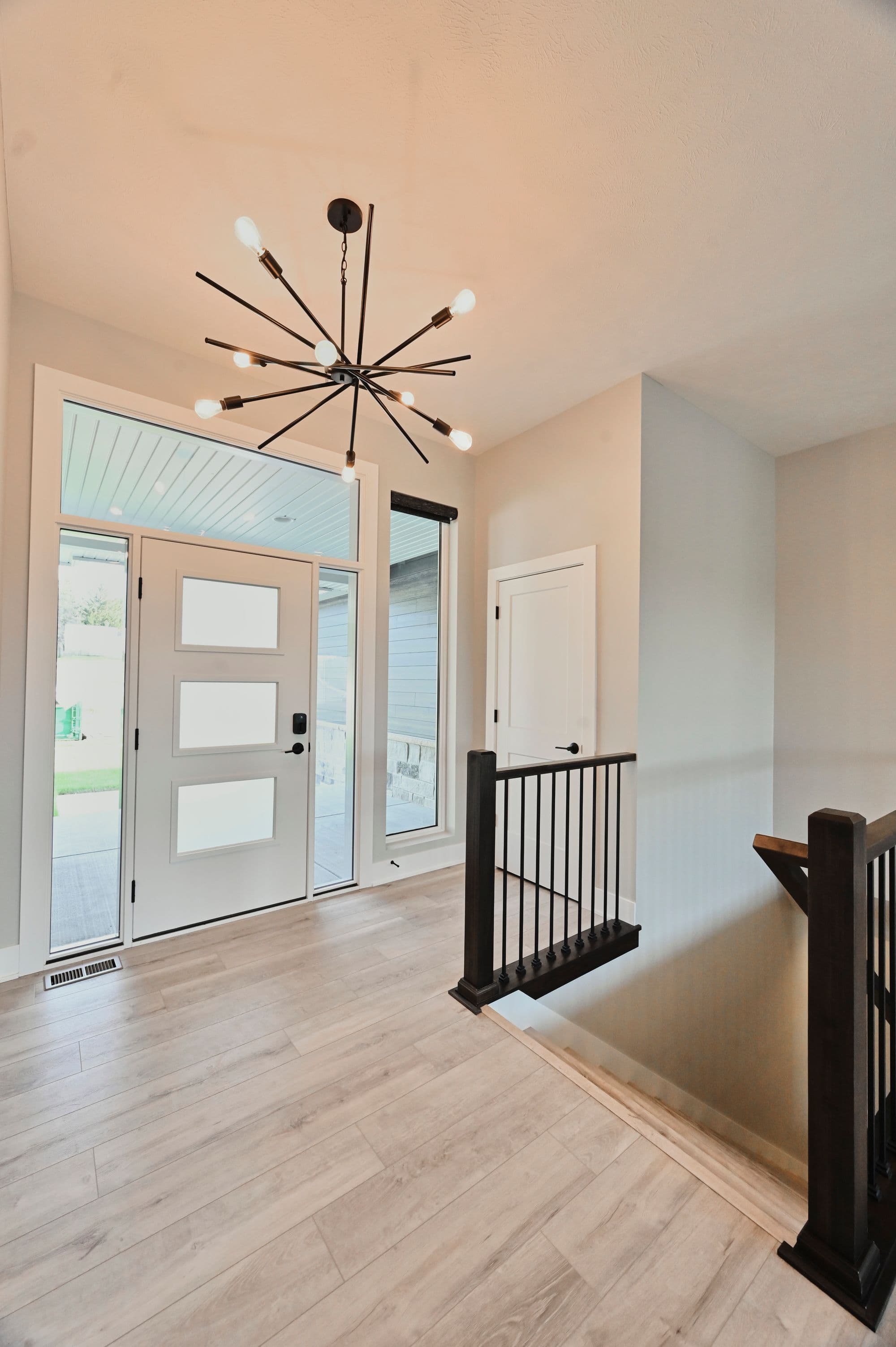 Modern home entry with light wood-look flooring, a white front door with square windows, a modern sputnik chandelier, and black metal railings leading to the staircase. The space is light and airy, with a view to the outside through the front door.