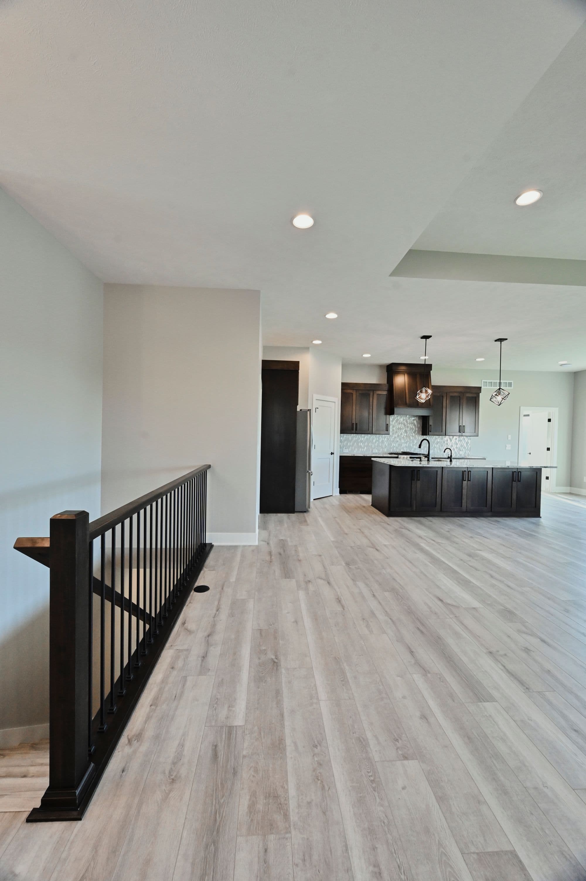 Open floor plan showcasing a modern kitchen with dark wood cabinetry and a large center island, connected to a living space with light gray flooring and a dark wood staircase railing. The kitchen features stainless steel appliances and pendant lighting.