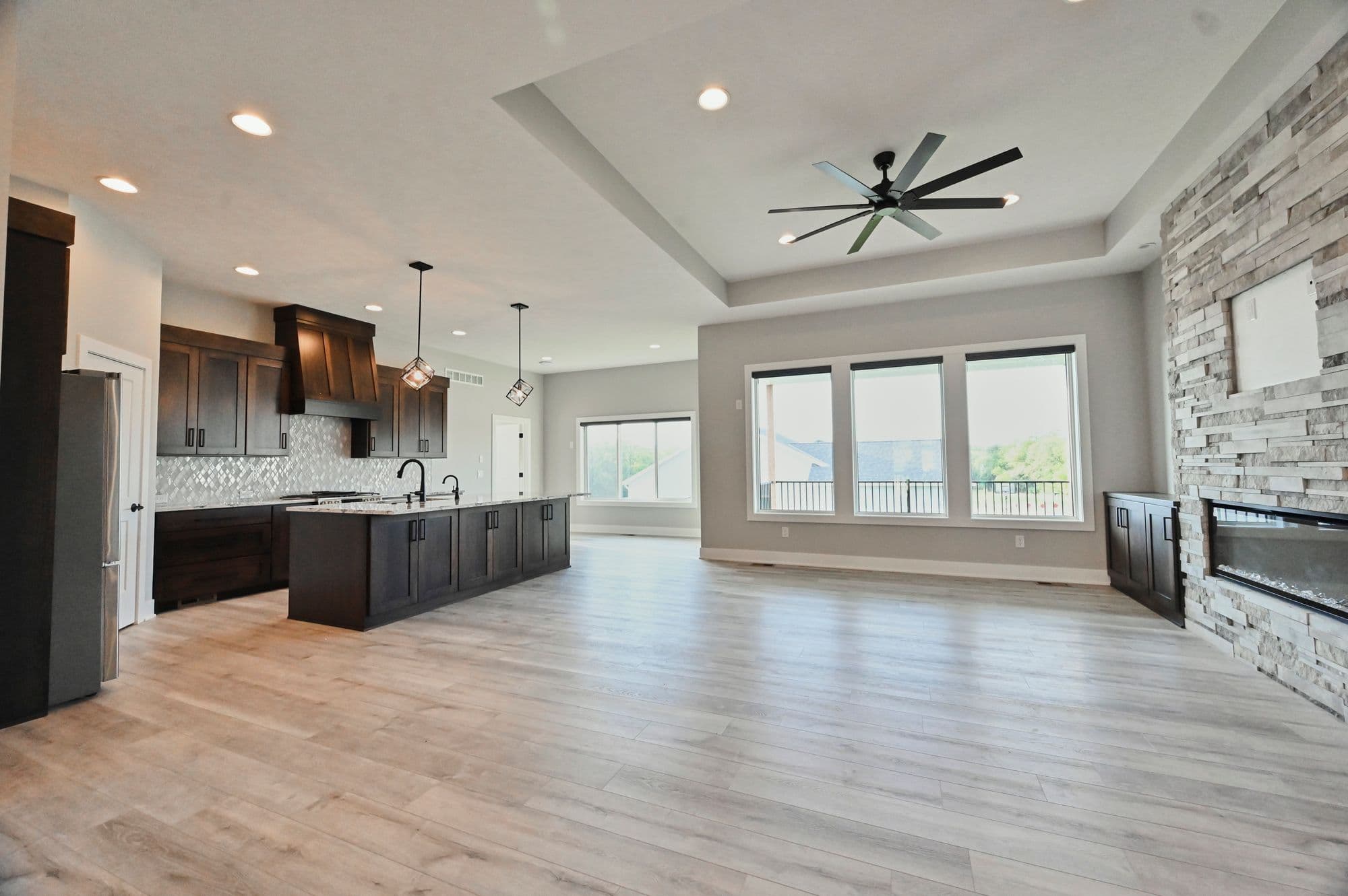 Open concept living space features a modern kitchen with dark wood cabinetry, an island with pendant lighting, and a living room with a stone fireplace and large windows. Hardwood floors run throughout the space, creating a cohesive design.
