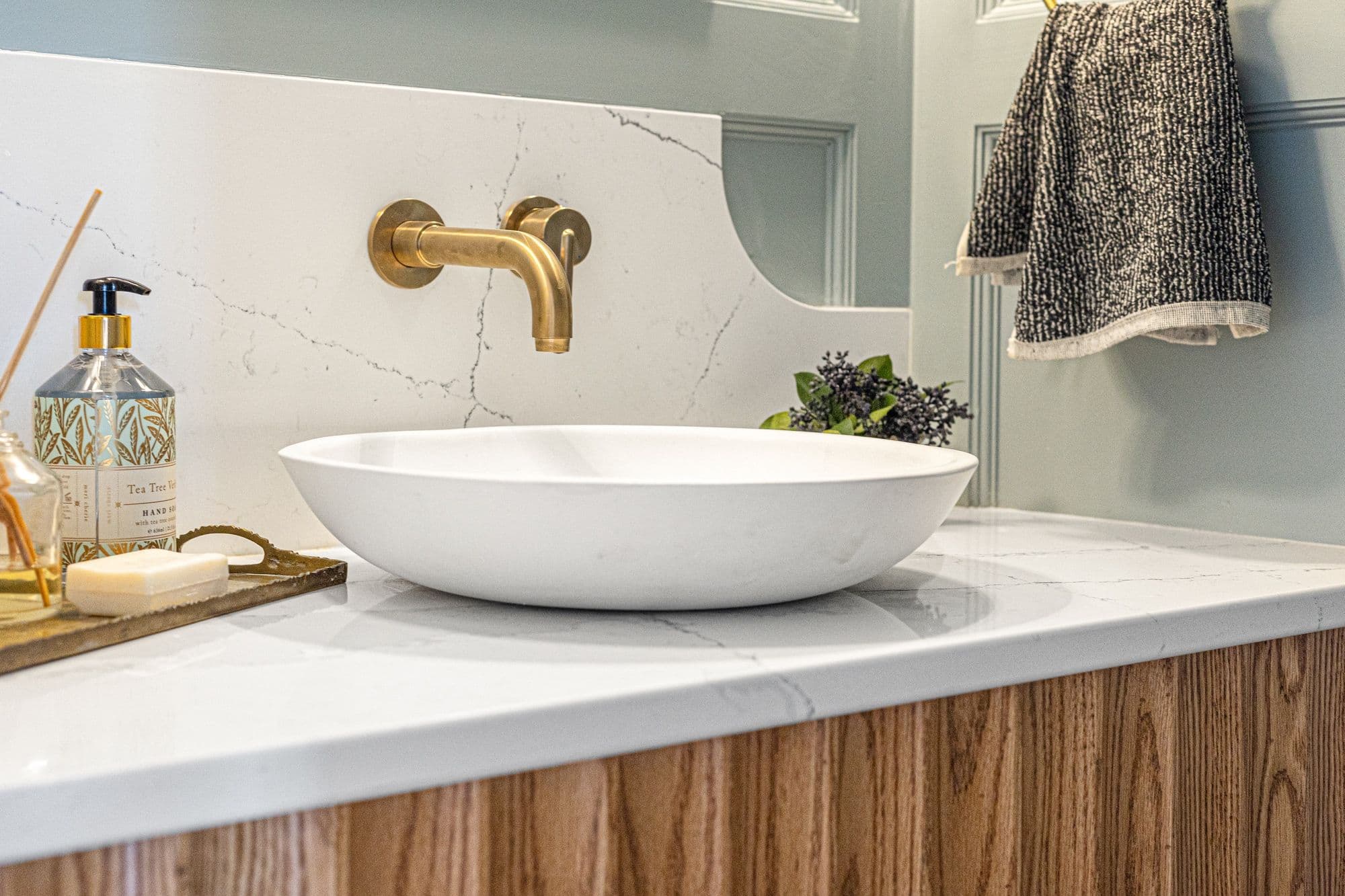 Bathroom vanity featuring a white countertop with marble-like veining, an oval-shaped vessel sink, and a wall-mounted gold faucet. The vanity is complemented by a light wood cabinet, and a textured hand towel hangs to the side, adding a decorative touch.
