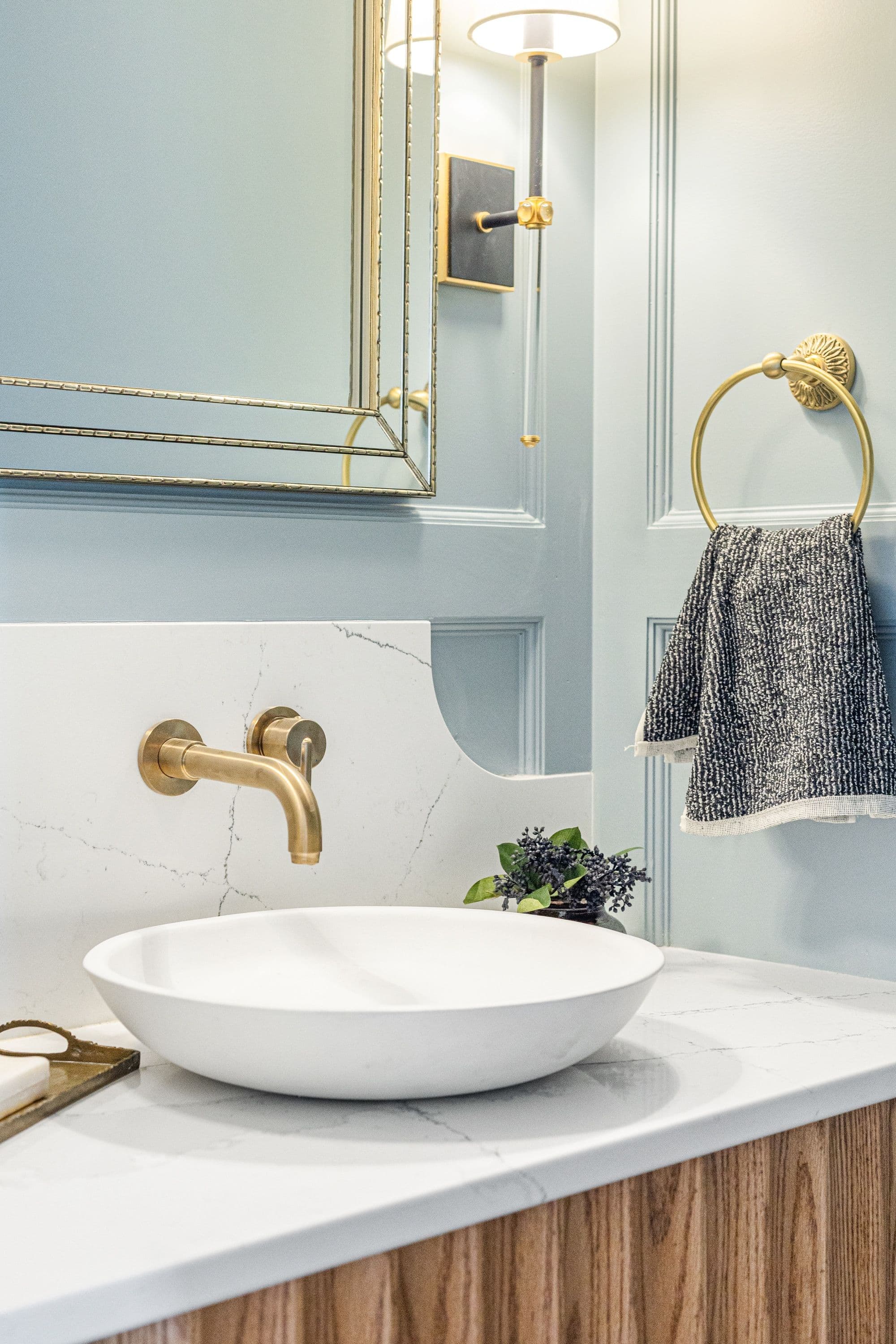 Modern bathroom vanity featuring a white vessel sink, brass faucet and towel ring, and blue-gray paneled walls. The countertop is white quartz with gray veining.