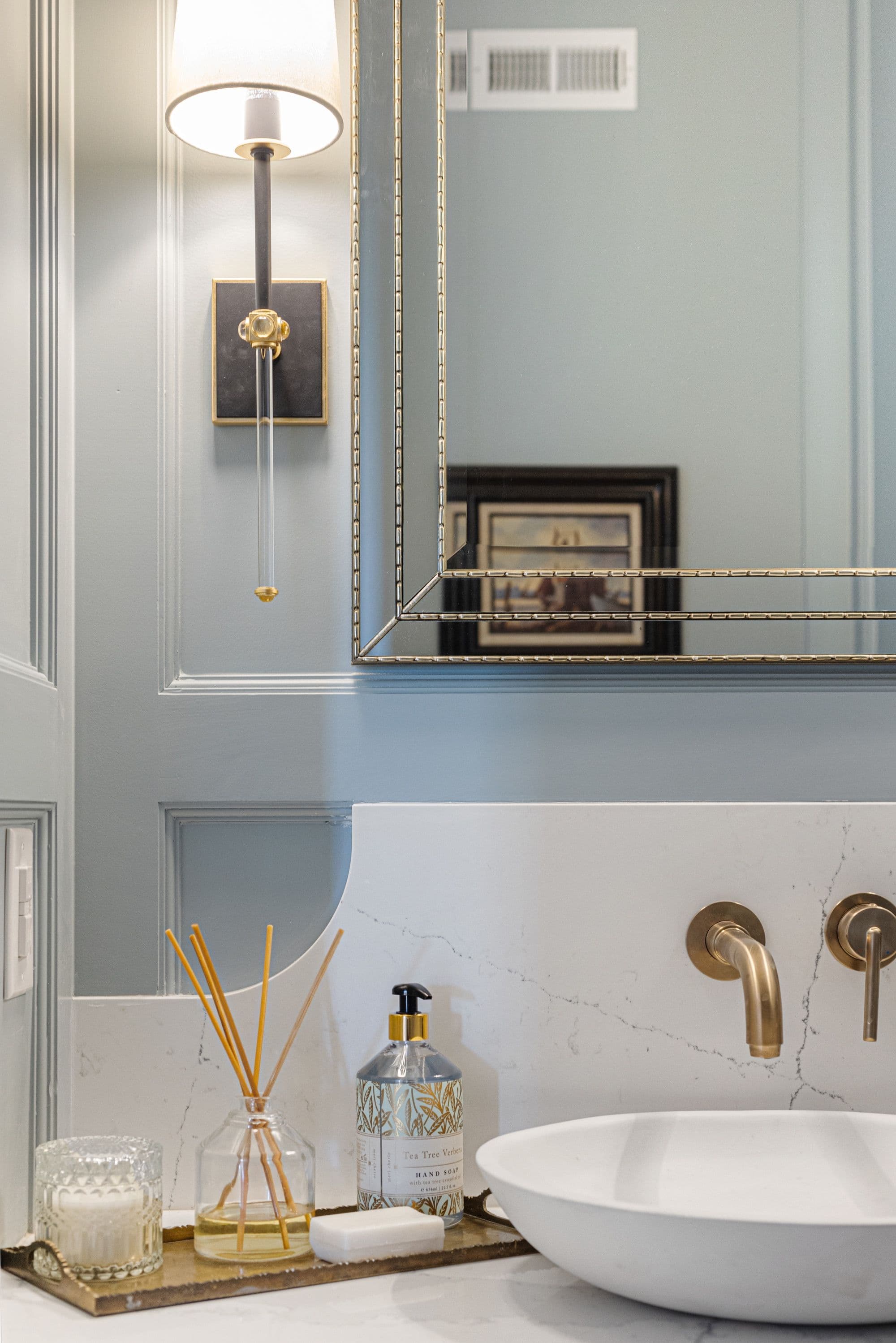 Bathroom vanity with a white marble countertop, white vessel sink, and brass fixtures is presented in this interior shot. A decorative mirror hangs above the vanity, reflecting artwork, while a sconce provides lighting.