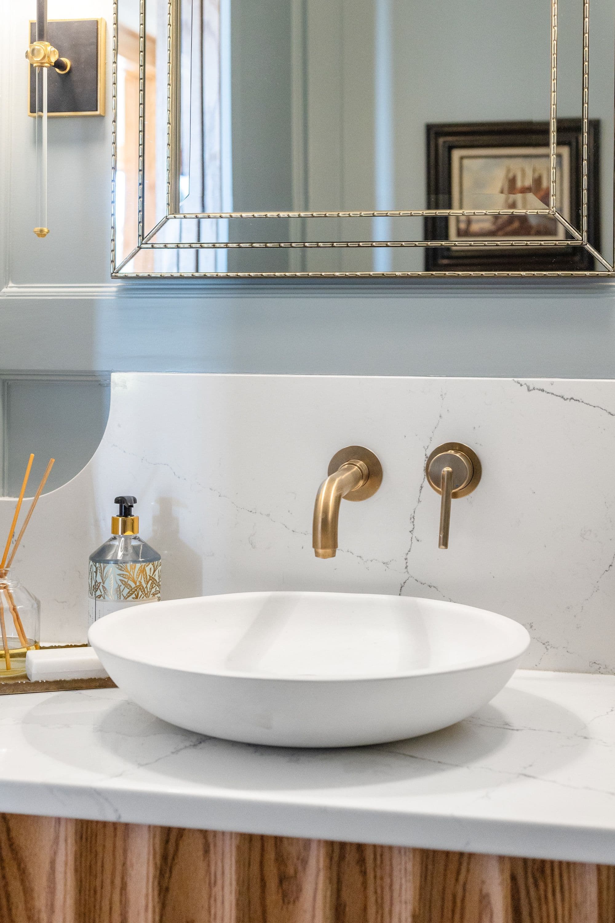 Bathroom vanity features a white vessel sink, marble countertop, and wall-mounted brass faucet. A framed mirror and decorative wall sconce accent the space, creating a luxurious and modern aesthetic.