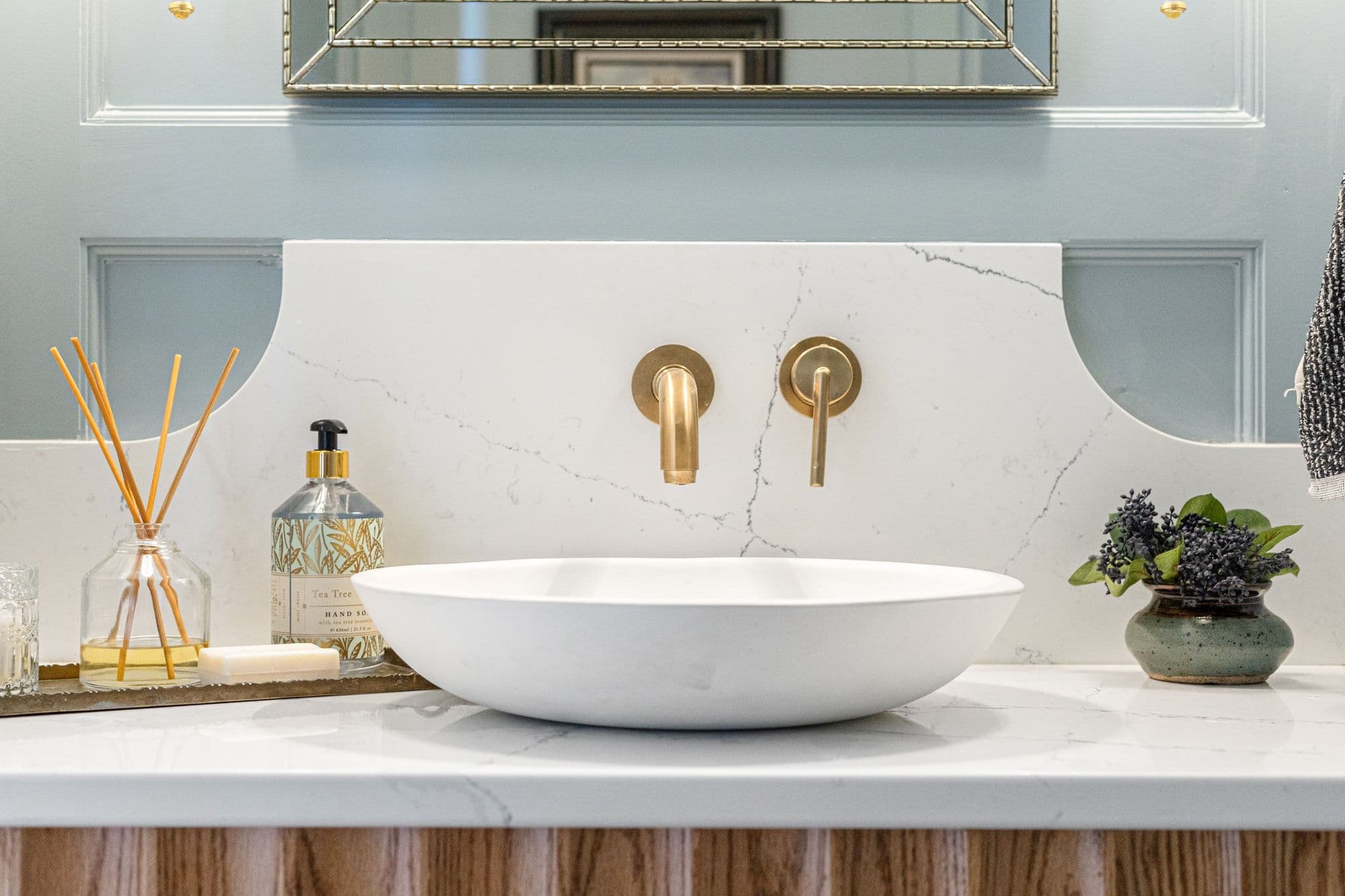 Modern bathroom features a white vessel sink, gold wall-mounted faucet, and marble backsplash. Decorative elements include a reed diffuser, hand soap, and a small vase with greenery.
