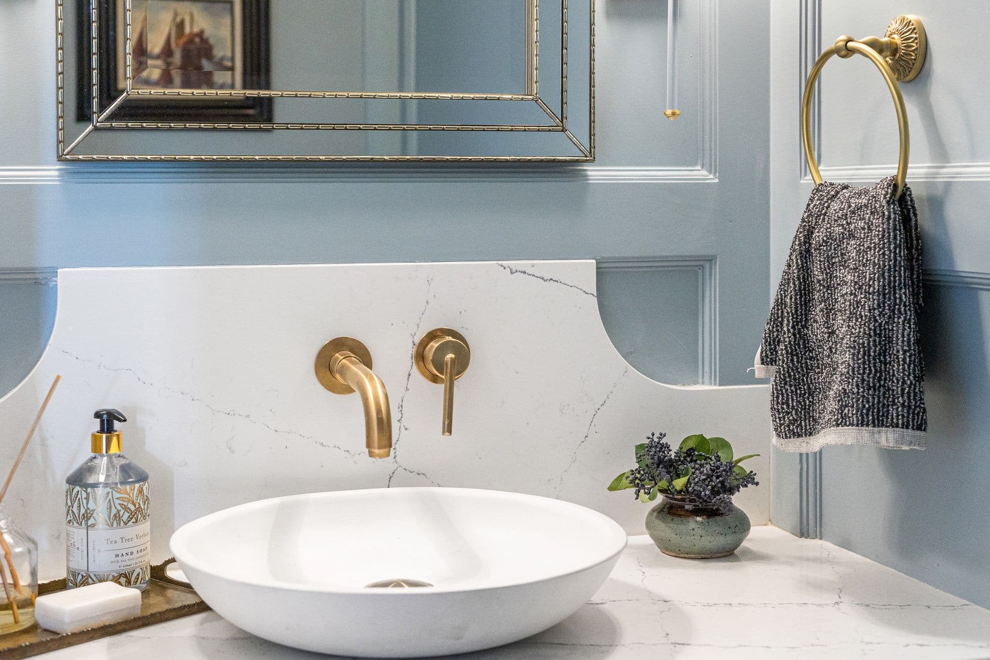 Bathroom vanity with a white vessel sink, gold faucet, and gray and white marble countertop. A round gold towel ring hangs on the blue paneled wall with a patterned hand towel.
