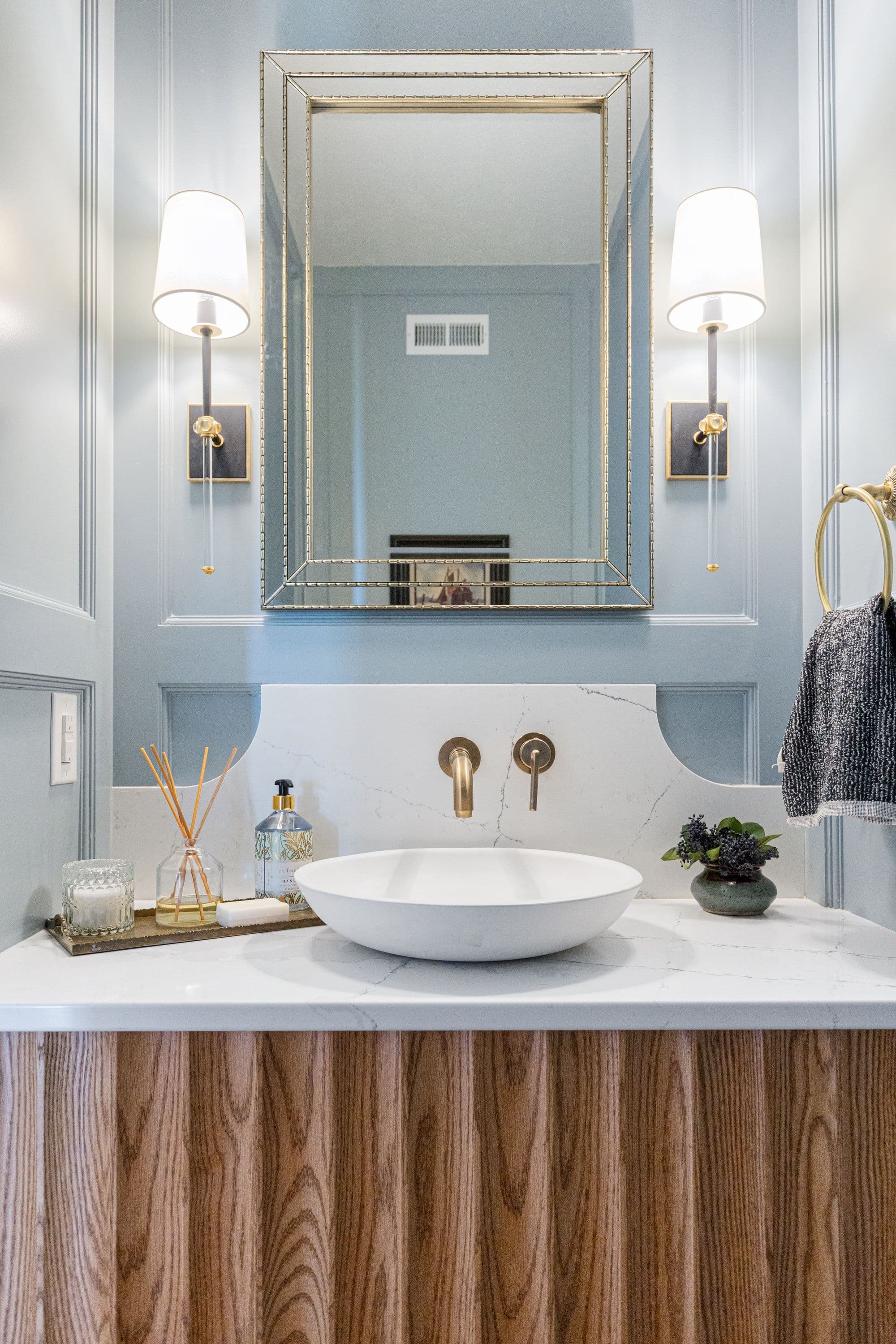 Powder room with vessel sink on marble countertop and wooden vanity, accented with modern sconces and framed mirror. The walls are painted a light blue-gray color with decorative molding.