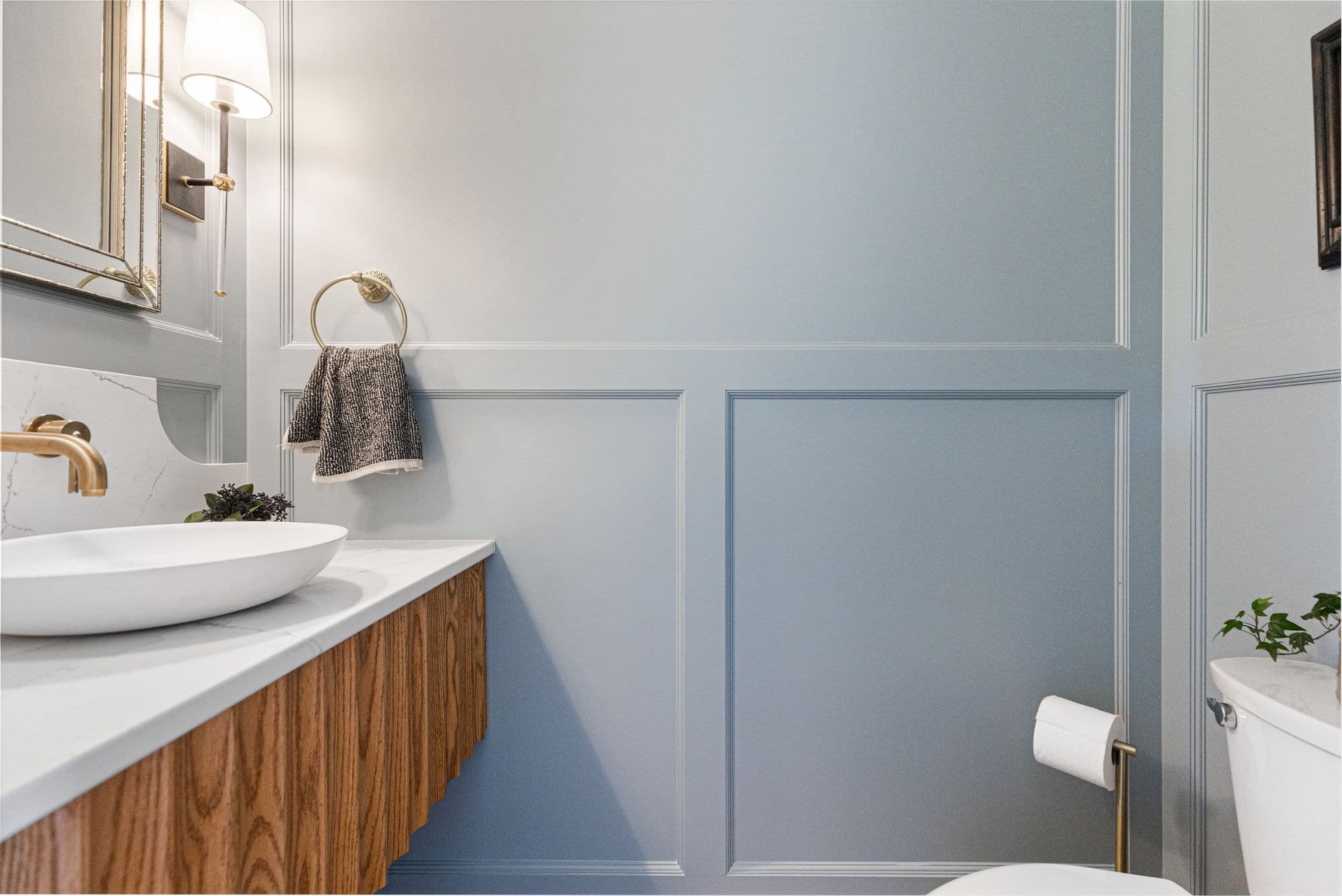 Powder room with blue paneled walls, a vessel sink, and a warm wood vanity. Gold fixtures accent the space, and a decorative mirror and sconce adorn the wall.