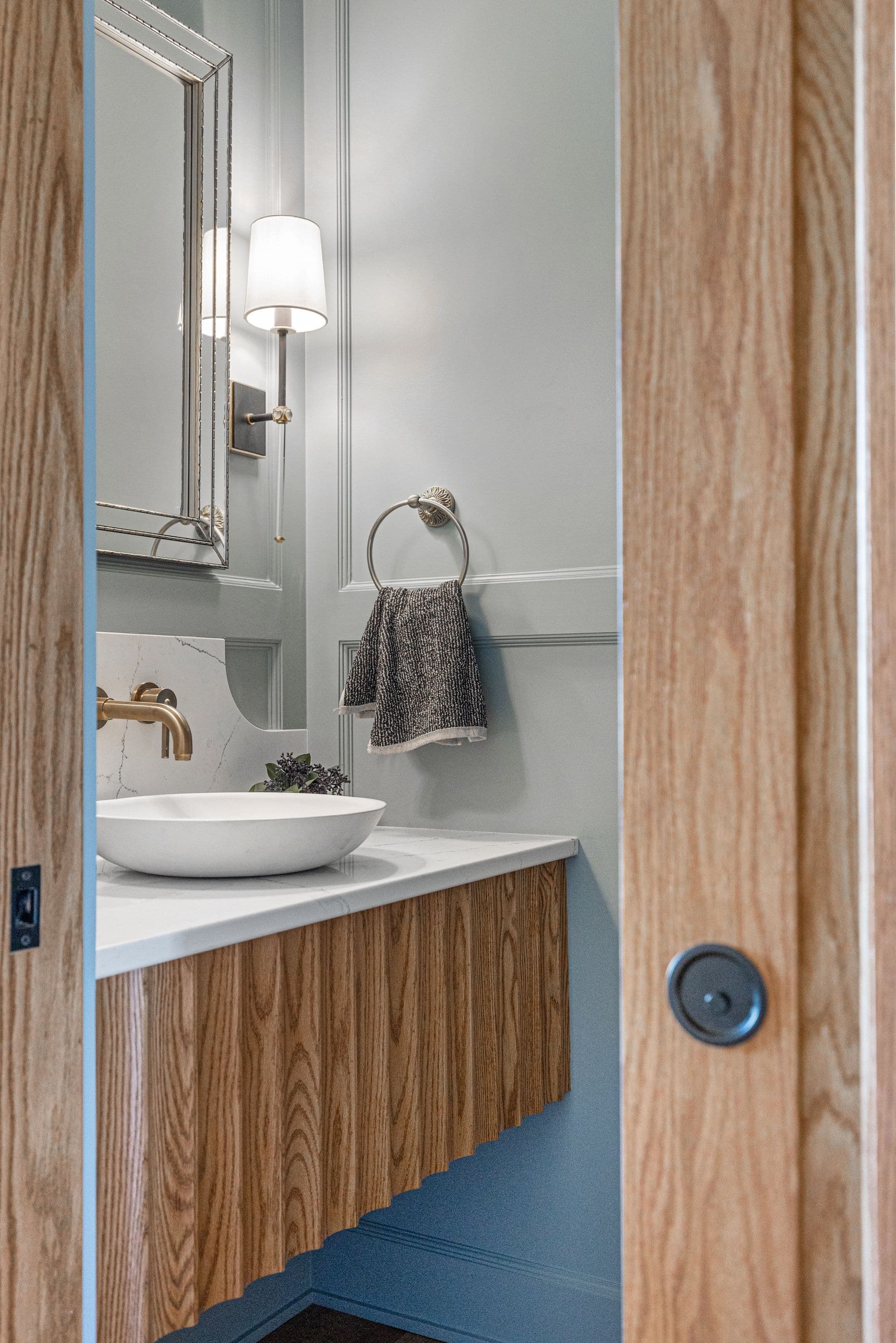 Modern bathroom featuring light gray walls, a wood-paneled vanity with a white countertop and vessel sink, and bronze fixtures. A silver-framed mirror and sconce flank the sink.