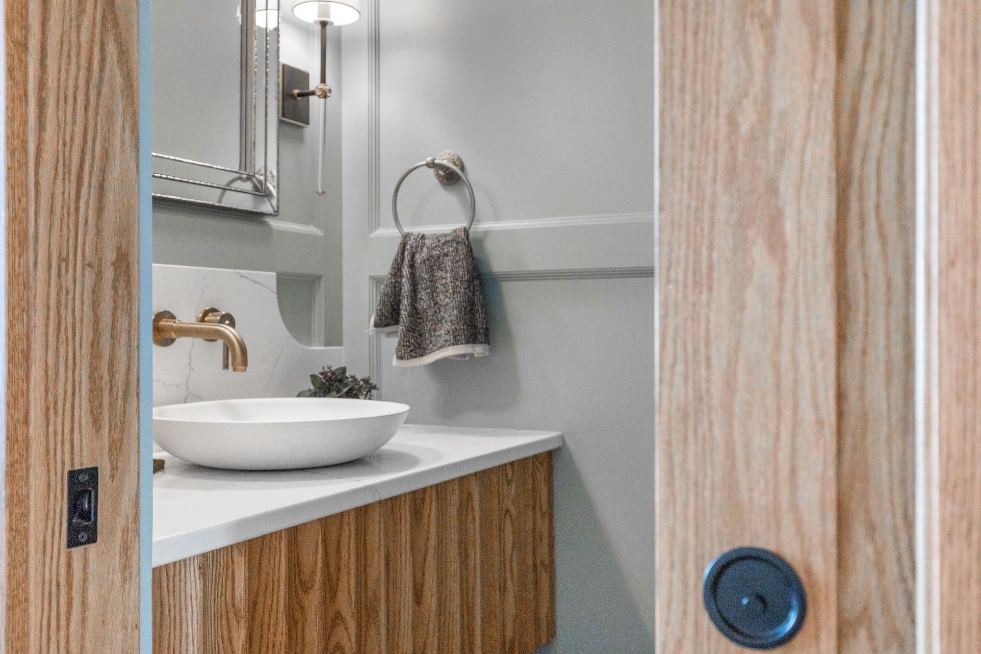 Bathroom view shows a round white sink with a gold faucet on a light wood vanity with a white countertop. A light gray textured towel hangs from a ring on the light gray wainscoted wall.