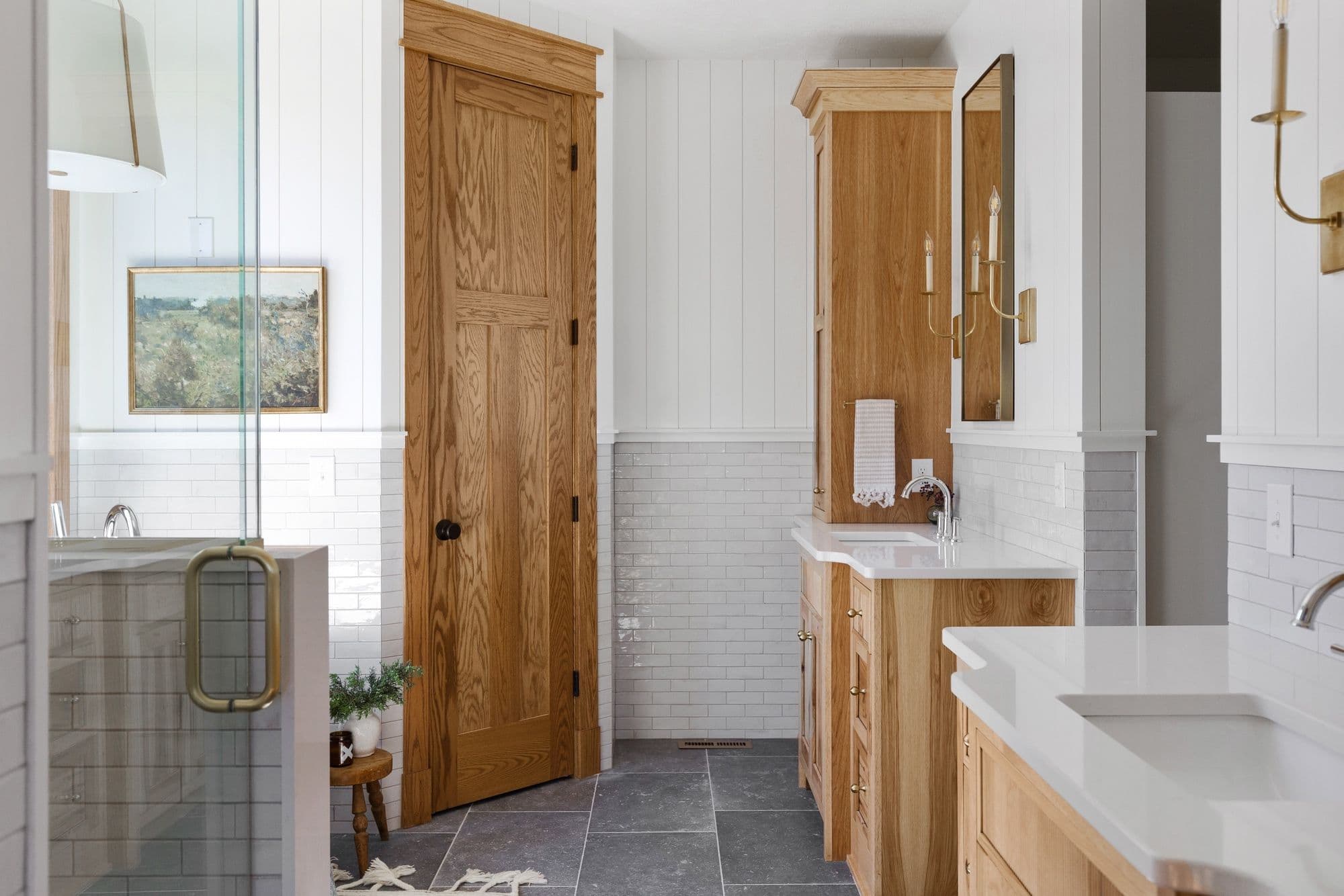 Traditional bathroom with oak vanity, subway tile wainscoting, and gray slate flooring. The room features a glass-enclosed shower, wood door, and brass sconces, creating a warm, inviting space.