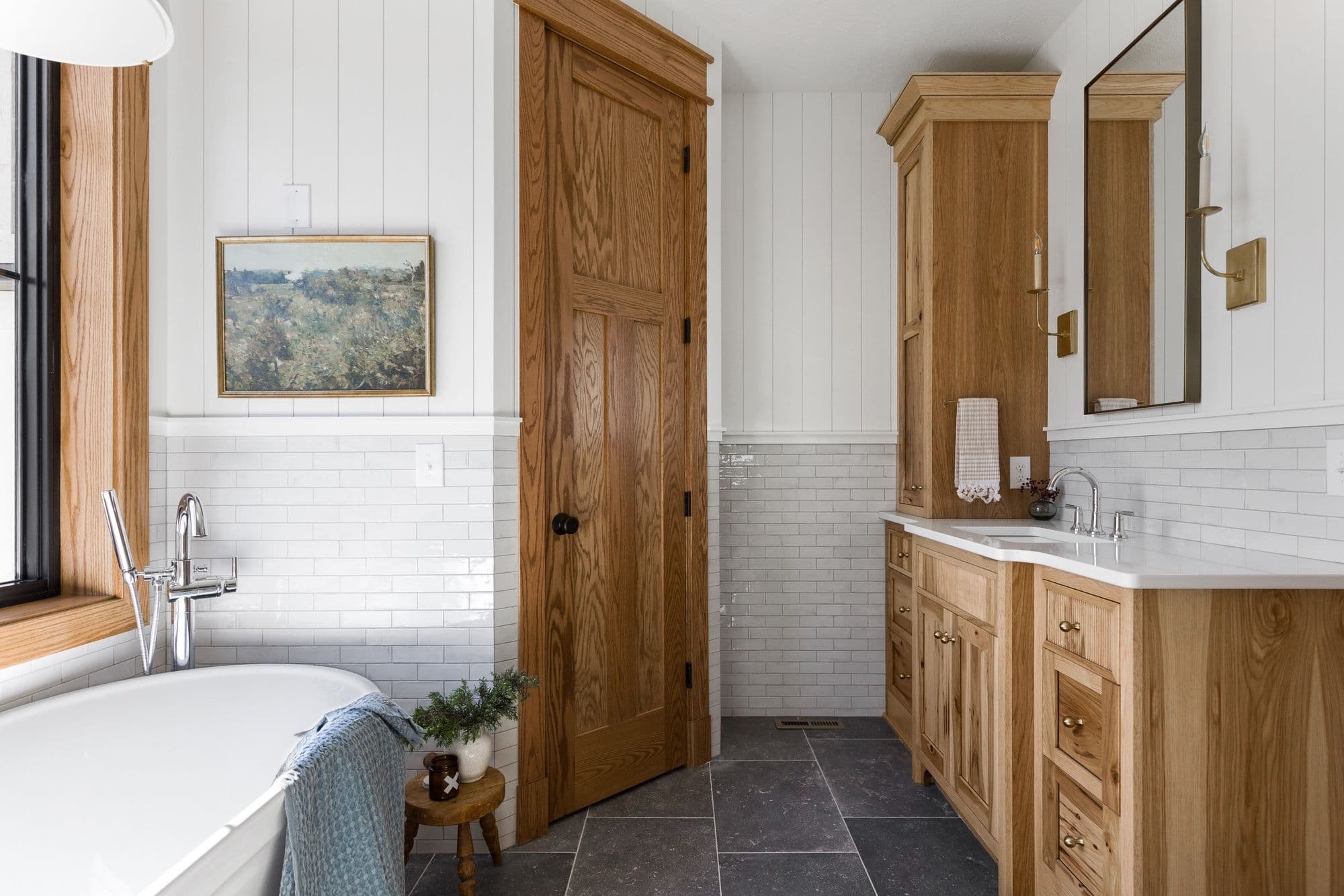 Bathroom interior featuring white subway tile, wood cabinets, and black tile flooring. The room is decorated with a framed landscape painting and has warm wood accents.