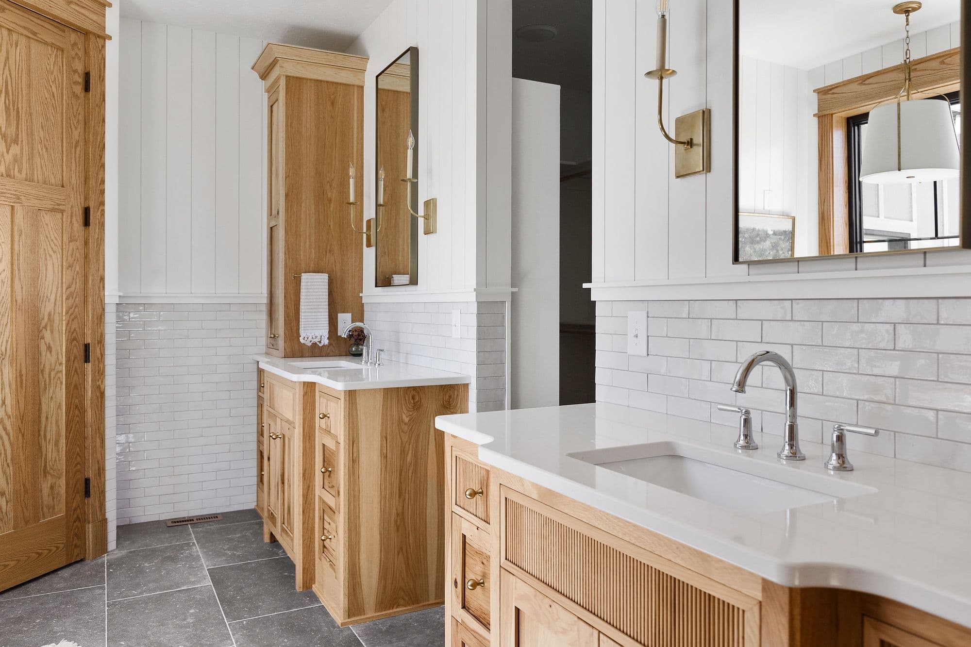 Well-lit bathroom featuring double vanities with natural wood cabinets, white quartz countertops, and subway tile backsplash. The space is accented with modern brass sconces and black-framed mirrors, complemented by light gray floor tiles and white plank walls.