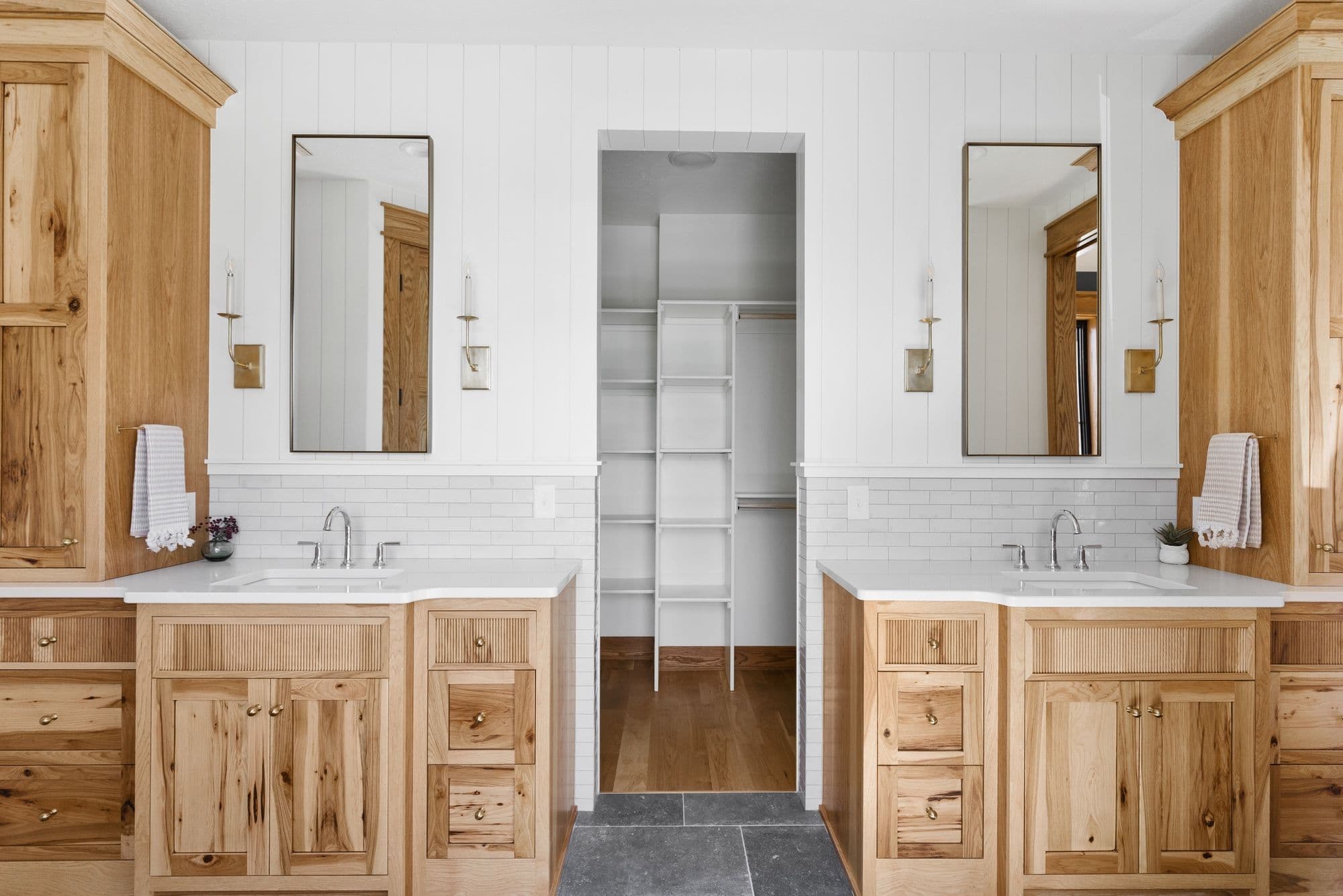 Bright and airy bathroom featuring double vanities crafted from warm wood tones, each accented with a rectangular mirror and sconces. The vanities are separated by an opening into a closet with white shelving and hardwood floors.