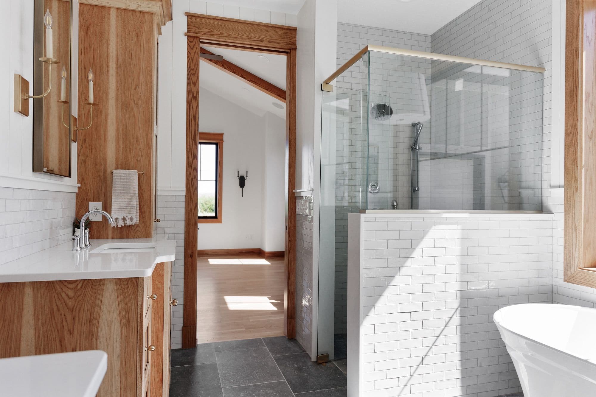 Bathroom featuring a wood vanity with a white countertop and a subway-tiled shower with glass enclosure. An open doorway leads into a room with wood floors and white walls under a vaulted ceiling.